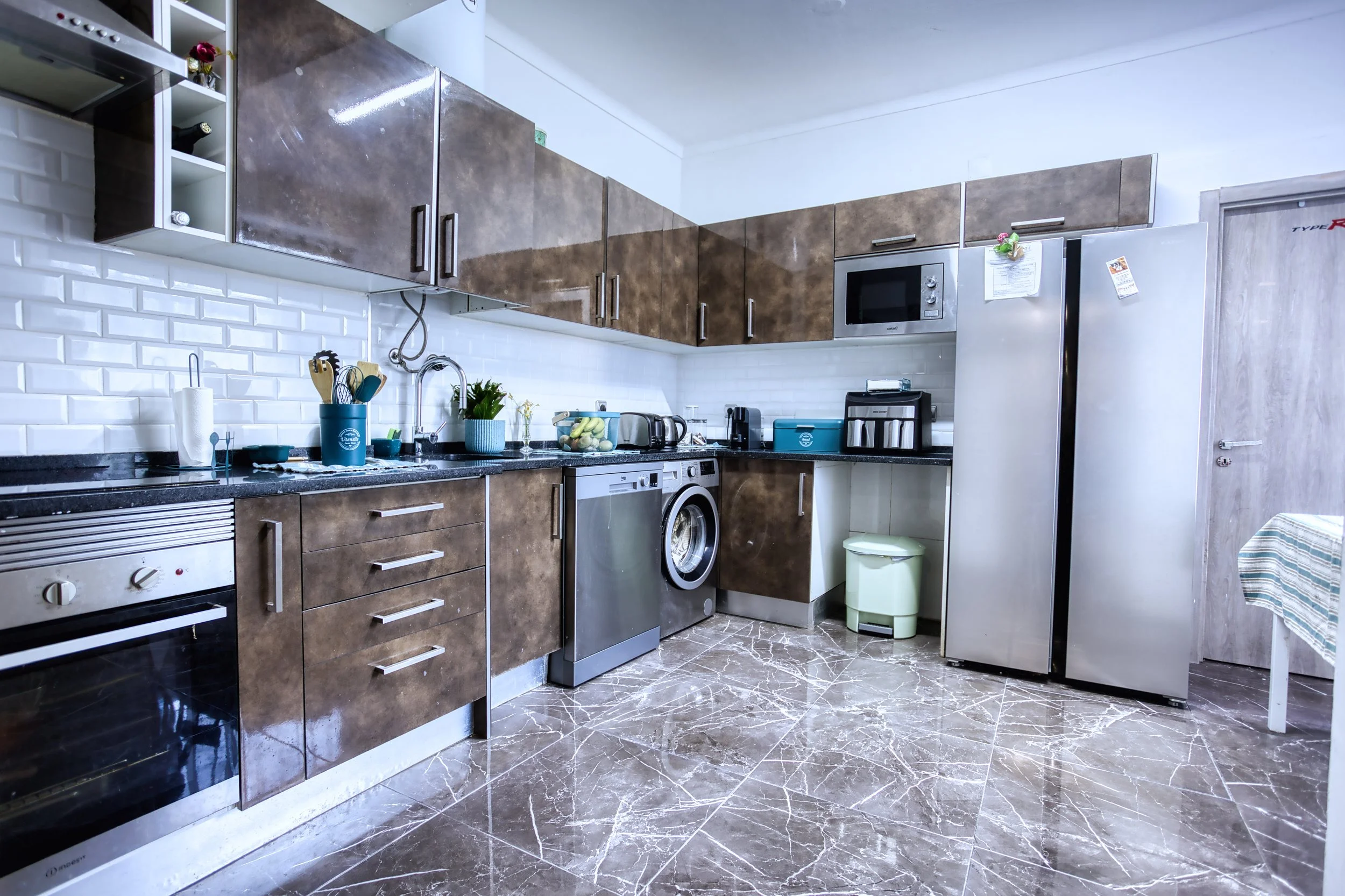 Cozinha moderna com armários marrons, bancada de granito preto, eletrodomésticos de aço inox, piso de mármore, parede de azulejos brancos, utensílios de cozinha e plantas decorativas.