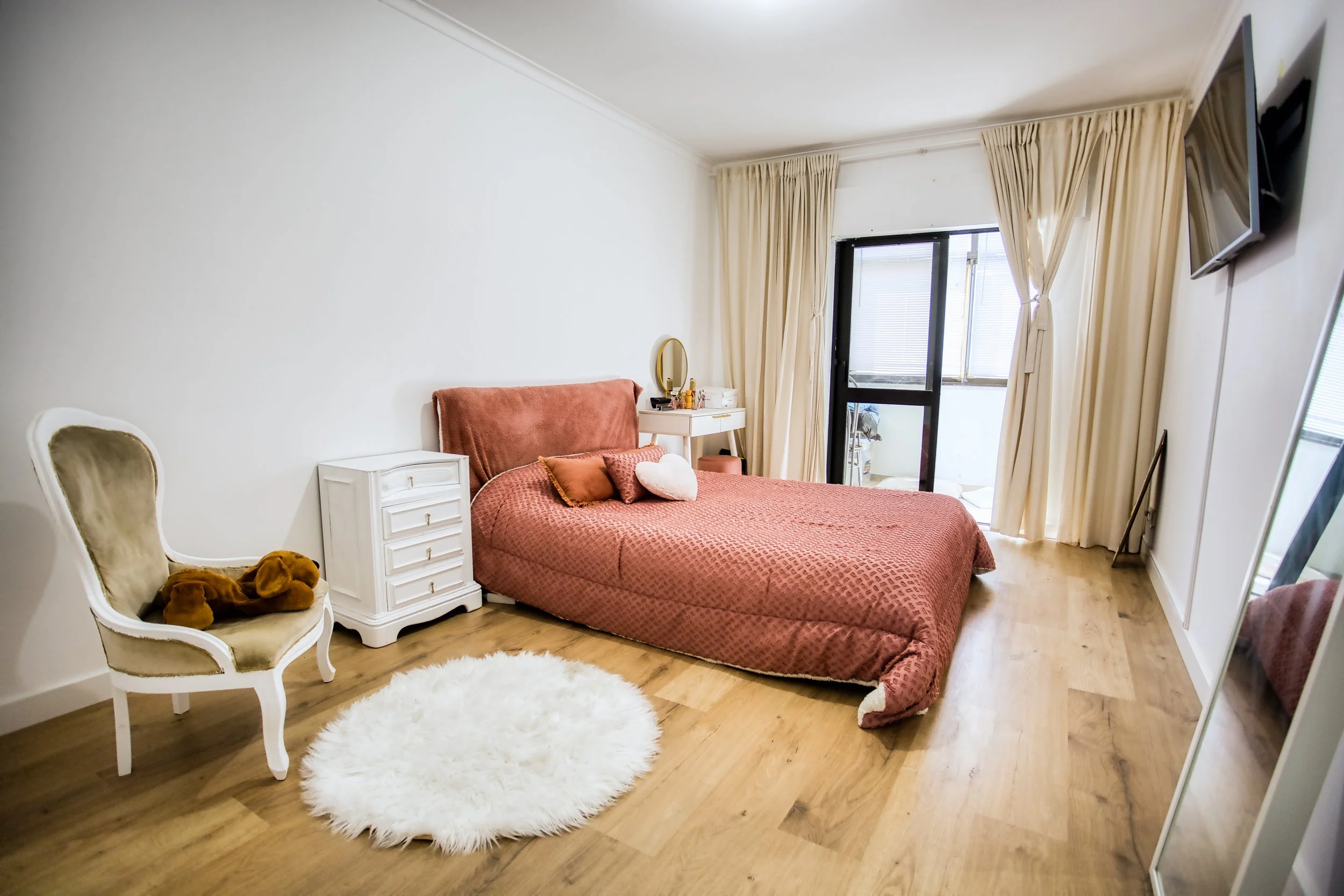 Quarto com cama de casal com cabeceira rosa, cortinas bege, televisão na parede, com uma cadeira de madeira e almofadas, espelho de corpo inteiro e tapete branco de pelos no chão de madeira.