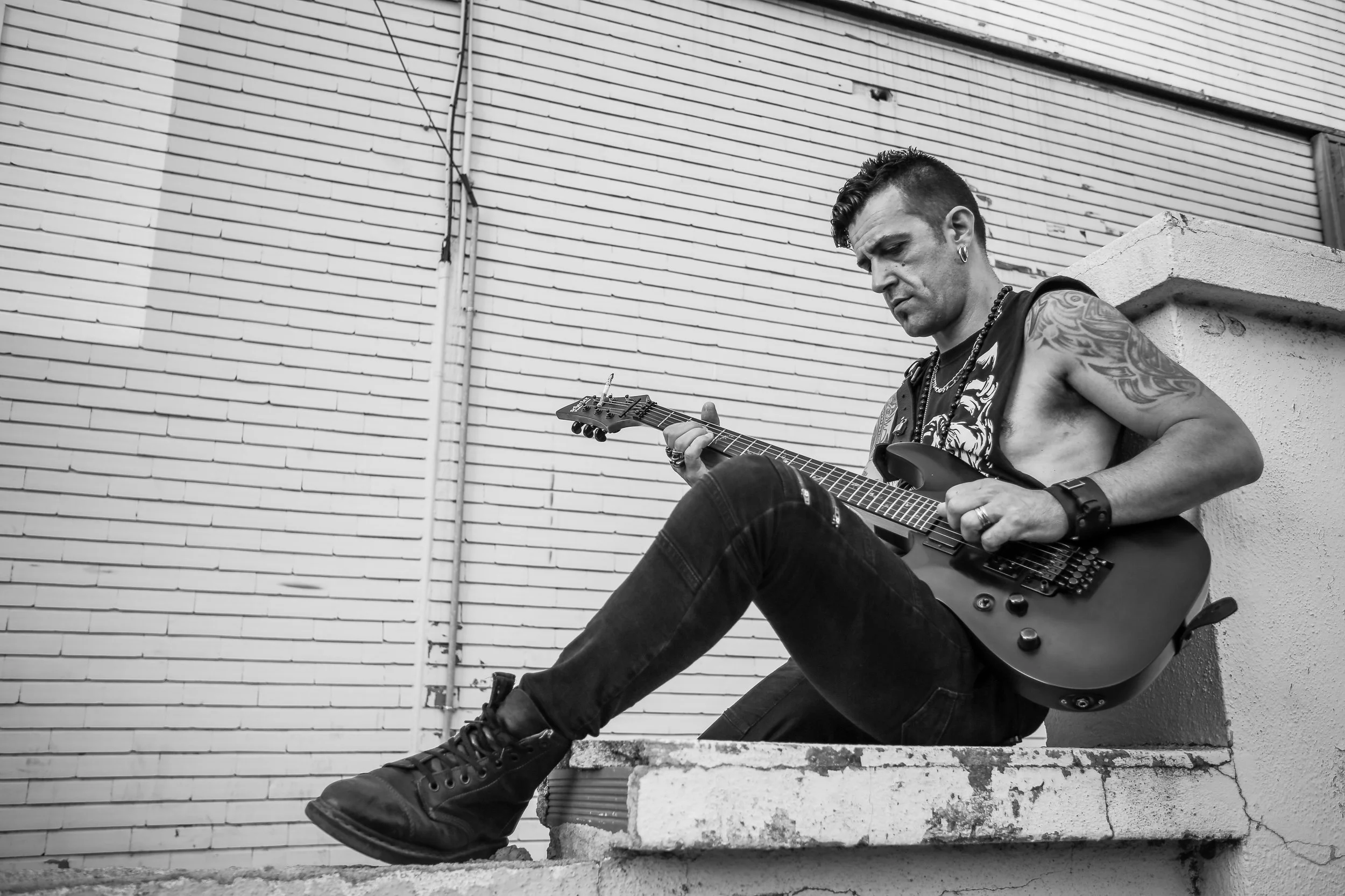 Homem tocando guitarra elétrica sentado em uma escada ao ar livre, com parede de tijolos ao fundo, em uma imagem em preto e branco.