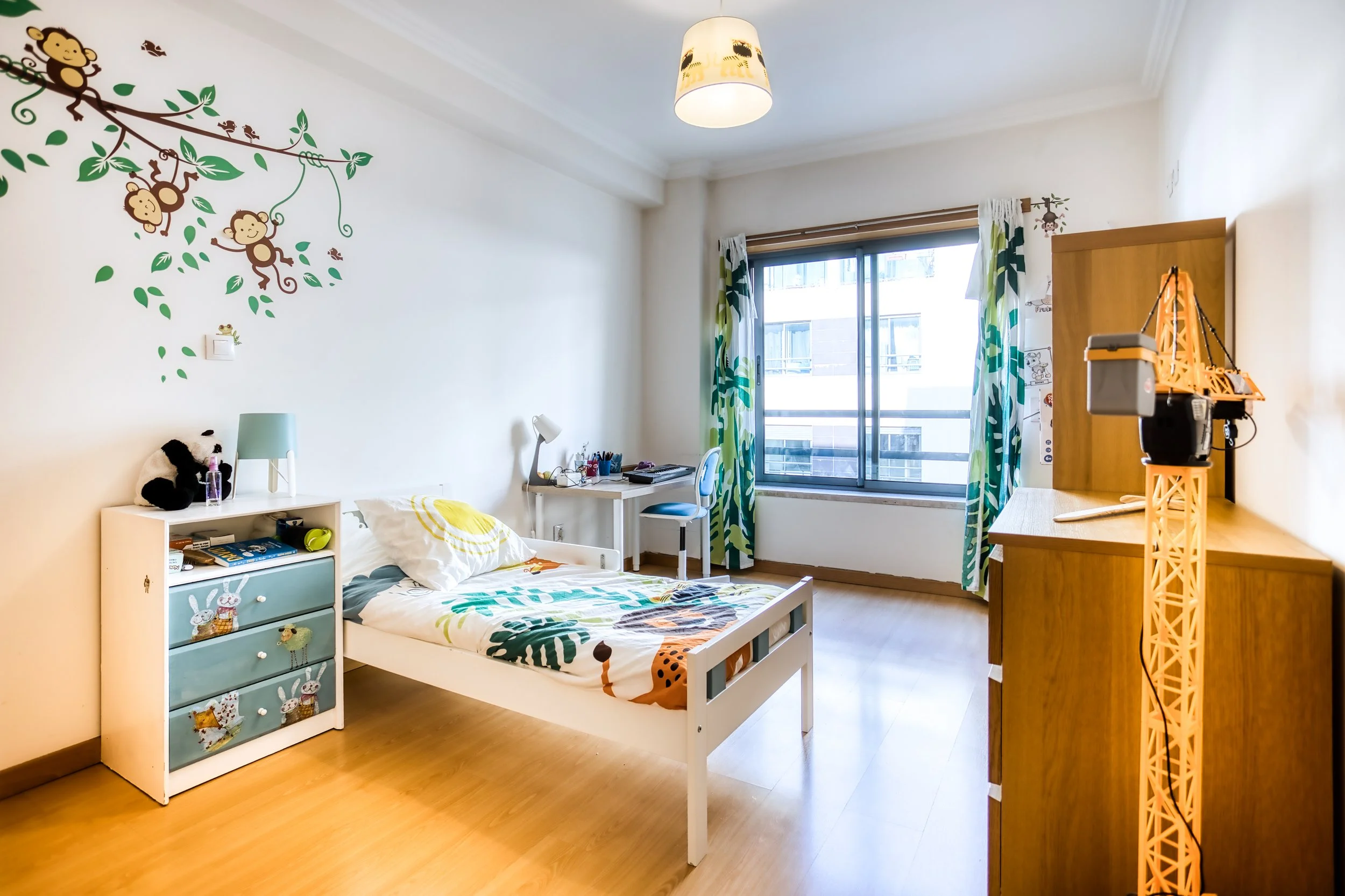 Quarto infantil com cama, armário de madeira, mesa de estudo, janela grande com cortinas de folhas verdes, parede decorada com desenhos de macacos e animais, piso de madeira, luminária no teto, brinquedo de construção em cima do armário.