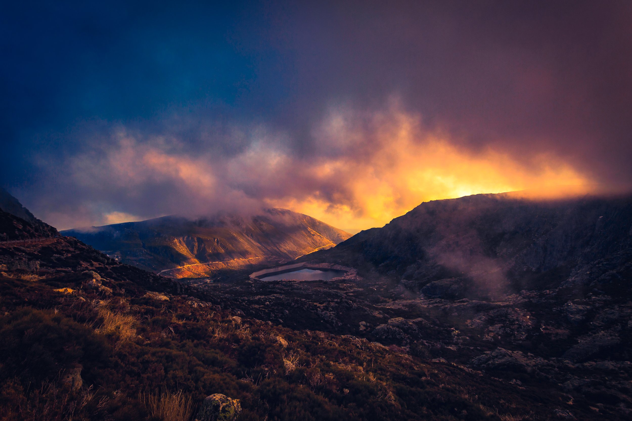 Cena de uma paisagem montanhosa com céu nublado ao entardecer, com árvores e um lago no vales, iluminados por luz dourada.