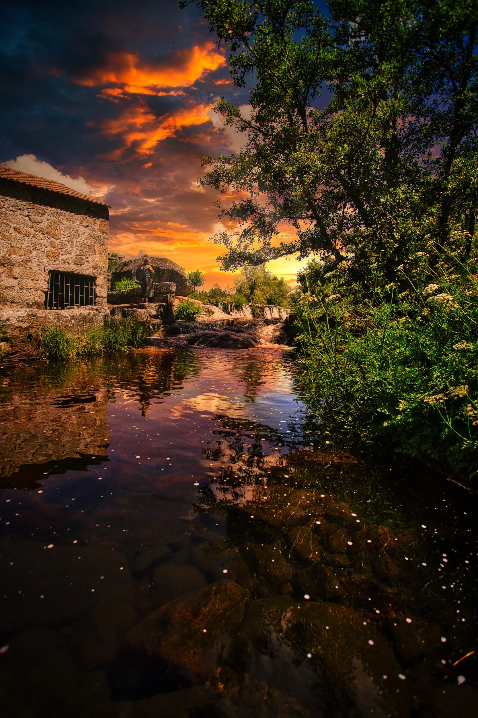 Céu ao pôr do sol com nuvens douradas e rosadas, árvores verdes à direita, uma casa de pedra ao lado de um riacho com pedras no fundo, refletindo o céu e a vegetação na água.