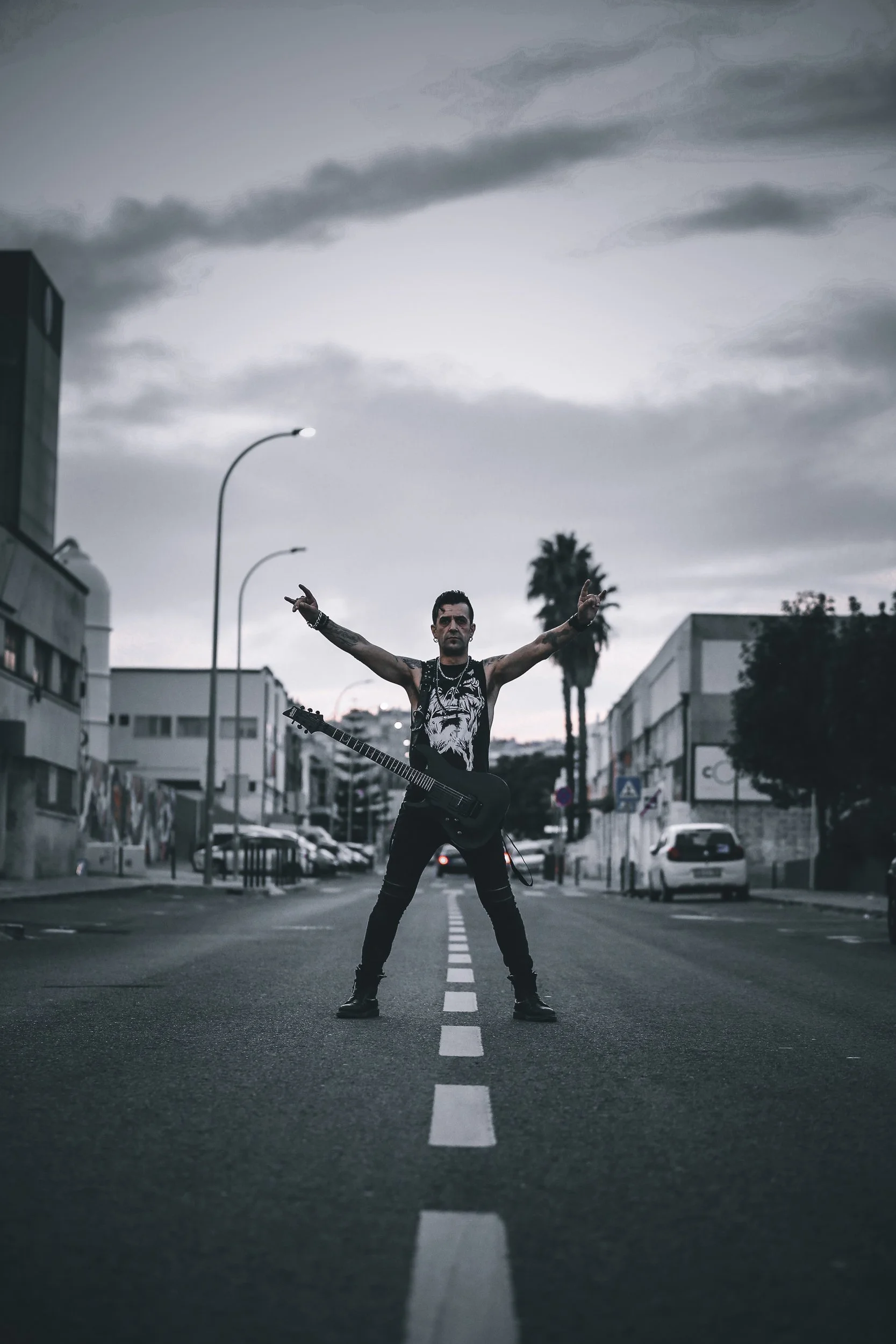 Homem parado no centro de uma rua de cidade, segurando uma guitarra elétrica, com os braços abertos e dedos formando o sinal de paz, ao entardecer sob céu nublado.