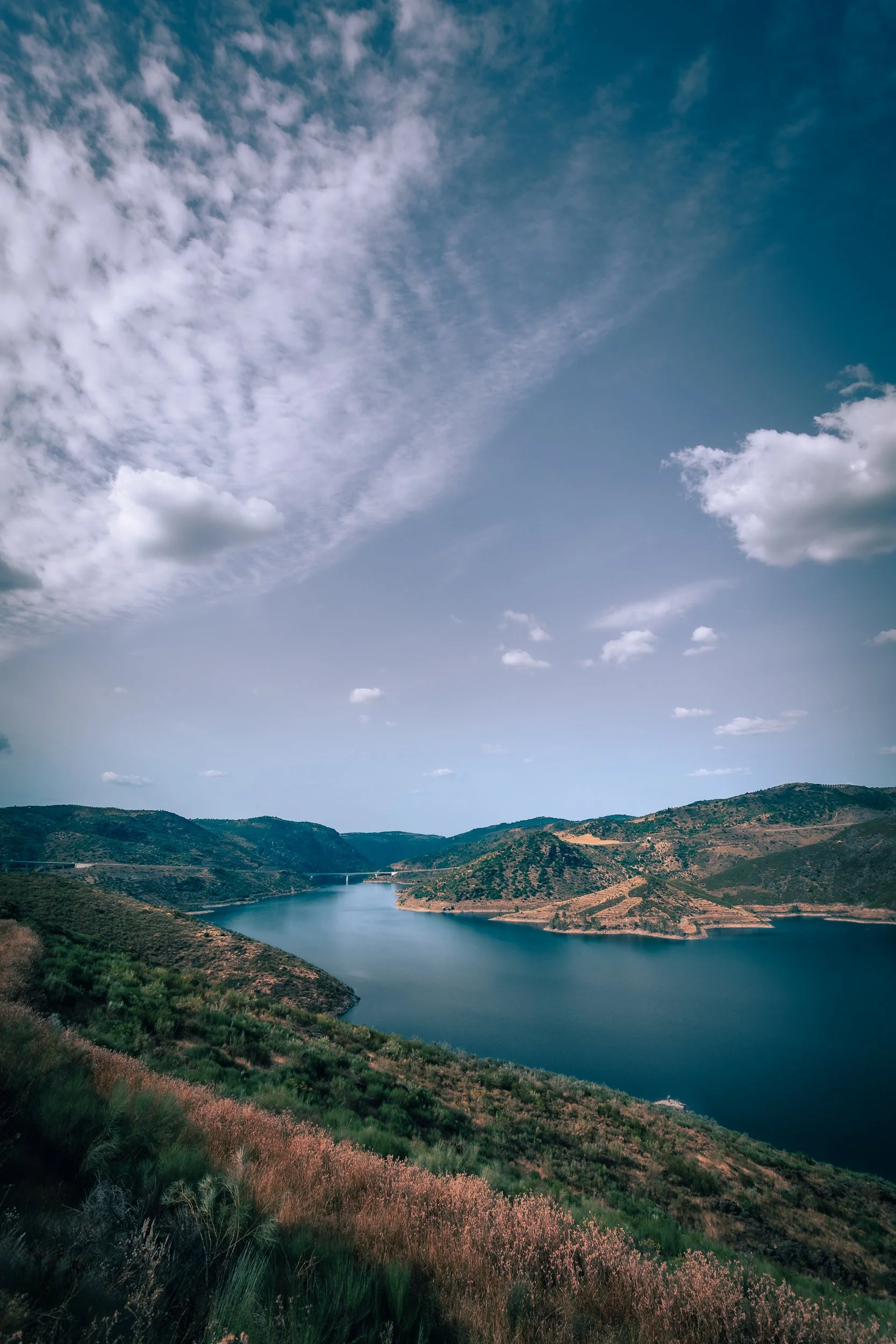 Lago com águas calmas cercado por colinas verdes e vegetação, céu claro com algumas nuvens.