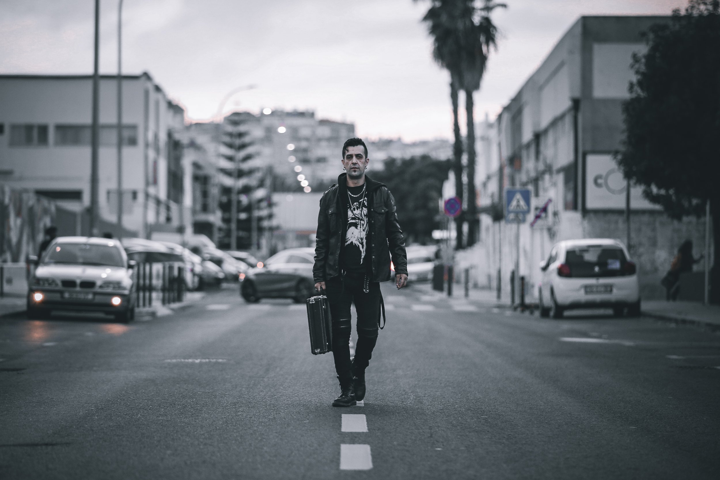 Homem andando no meio de uma rua urbana com malas, vestido com jaqueta de couro preto e roupas escuras, durante o pôr do sol