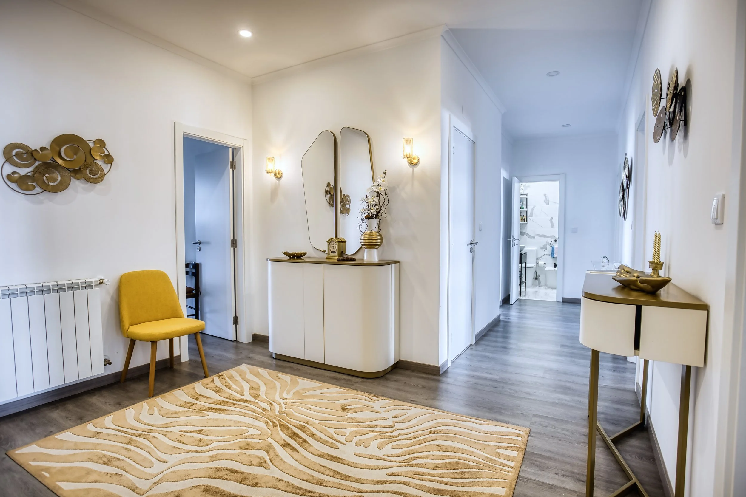 Sala de entrada decorada com móveis brancos, espelhos, luminárias de parede, uma cadeira amarela, uma bancada decorativa com vasos e objetos dourados, tapete claro com padrão de listras, paredes brancas e piso de madeira.