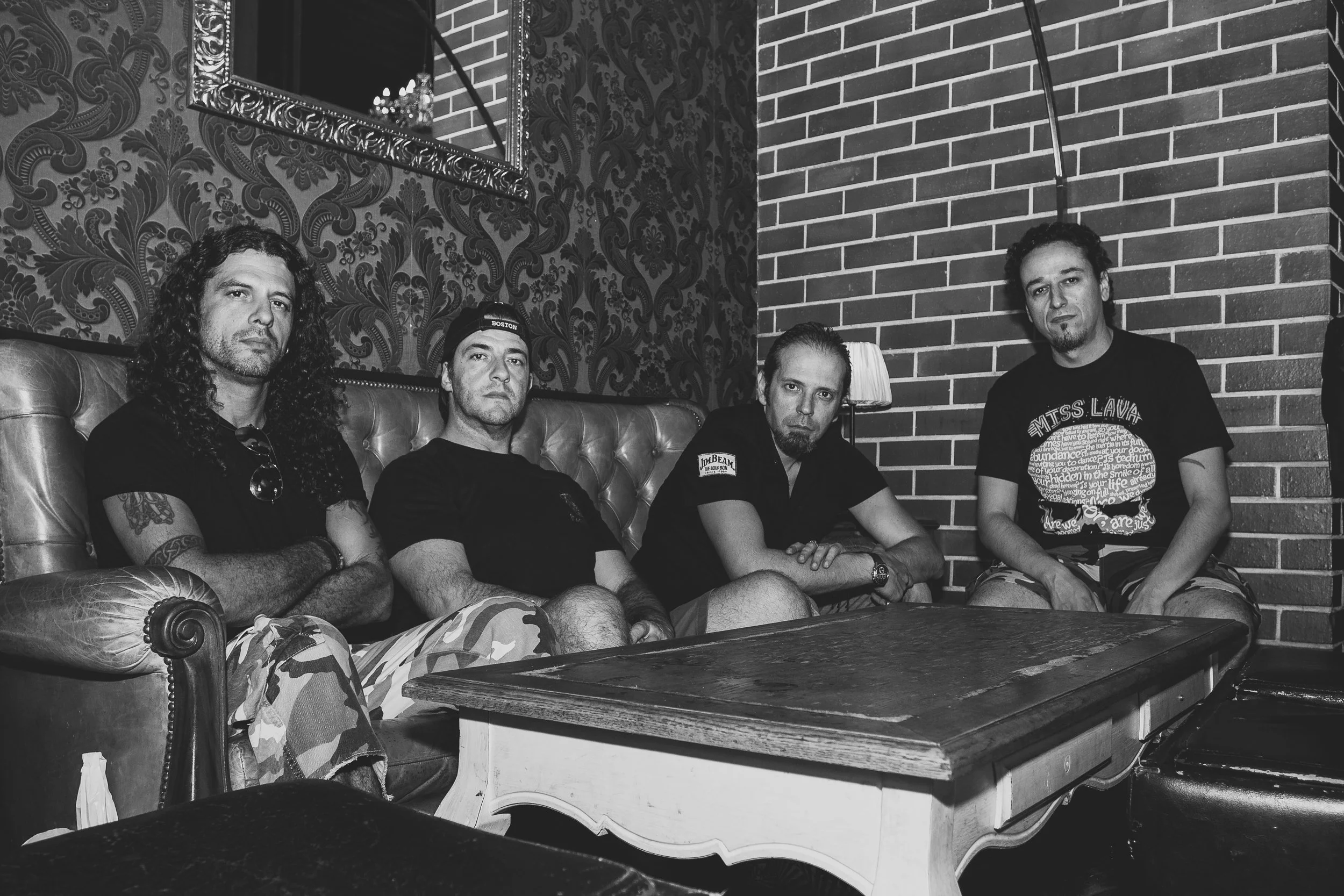 Quatro homens sentados em um sofá no interior de um ambiente, com paredes de tijolos e papel de parede com padrão floral escuro.