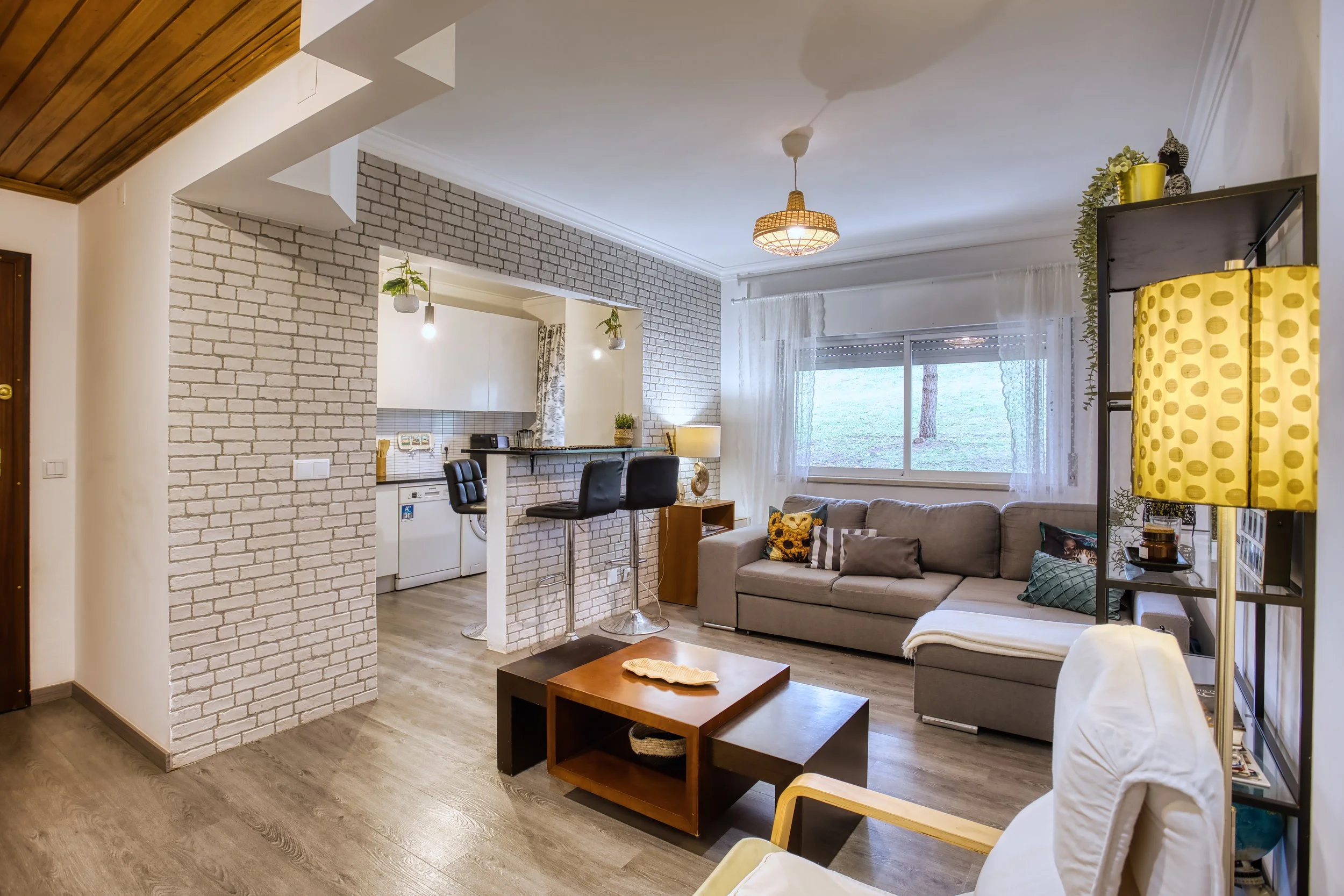 Sala de estar com sofá cinza, voltagem de janelas com cortinas brancas, luminária de piso amarela, mesa de centro de madeira, parede de tijolos brancos, bancada de cozinha com três cadeiras pretas e luzes penduradas, piso de madeira, decoração com pl