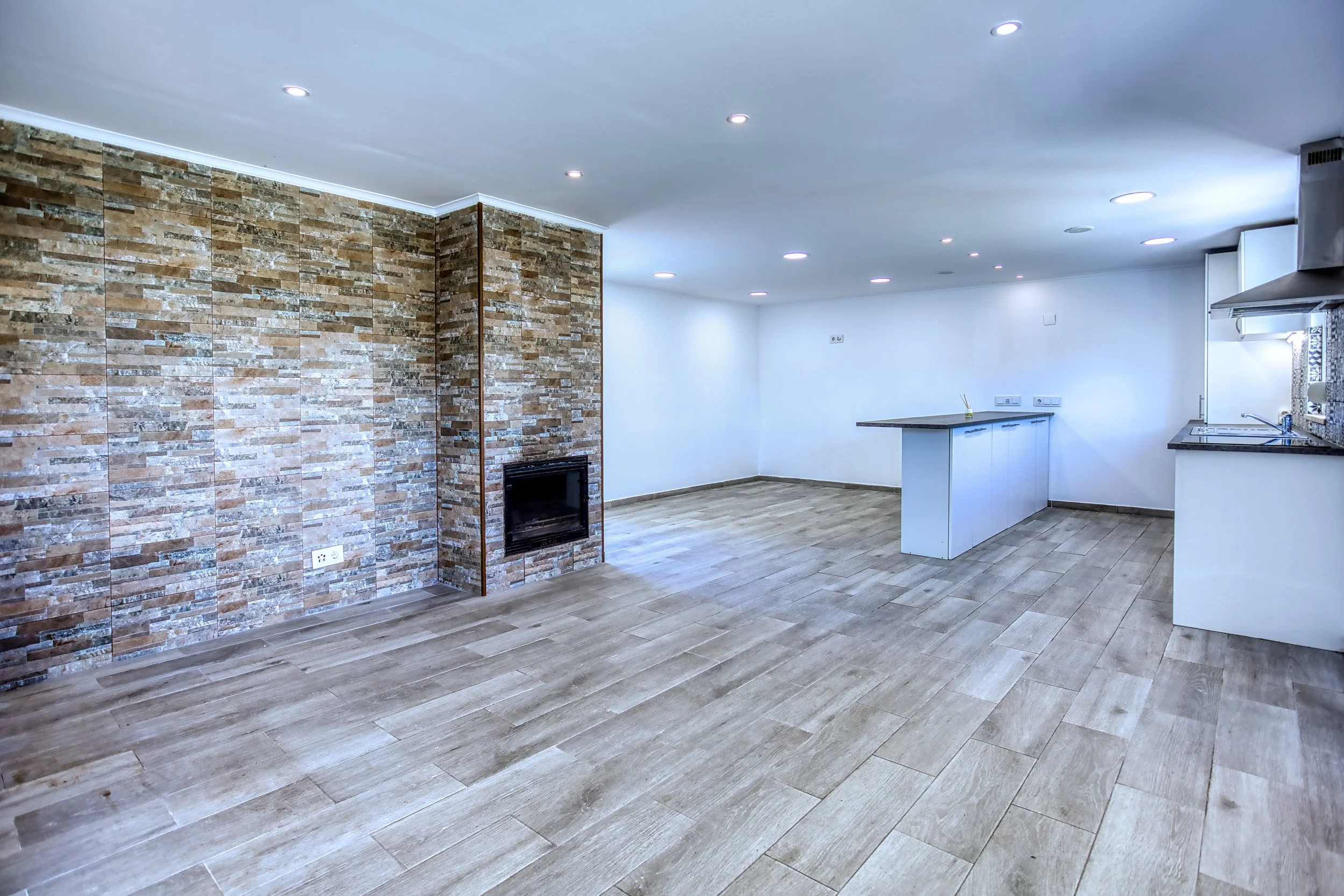 Sala de estar clara com piso de madeira, parede de pedra com lareira e cozinha integrada simples com bancada de granito.