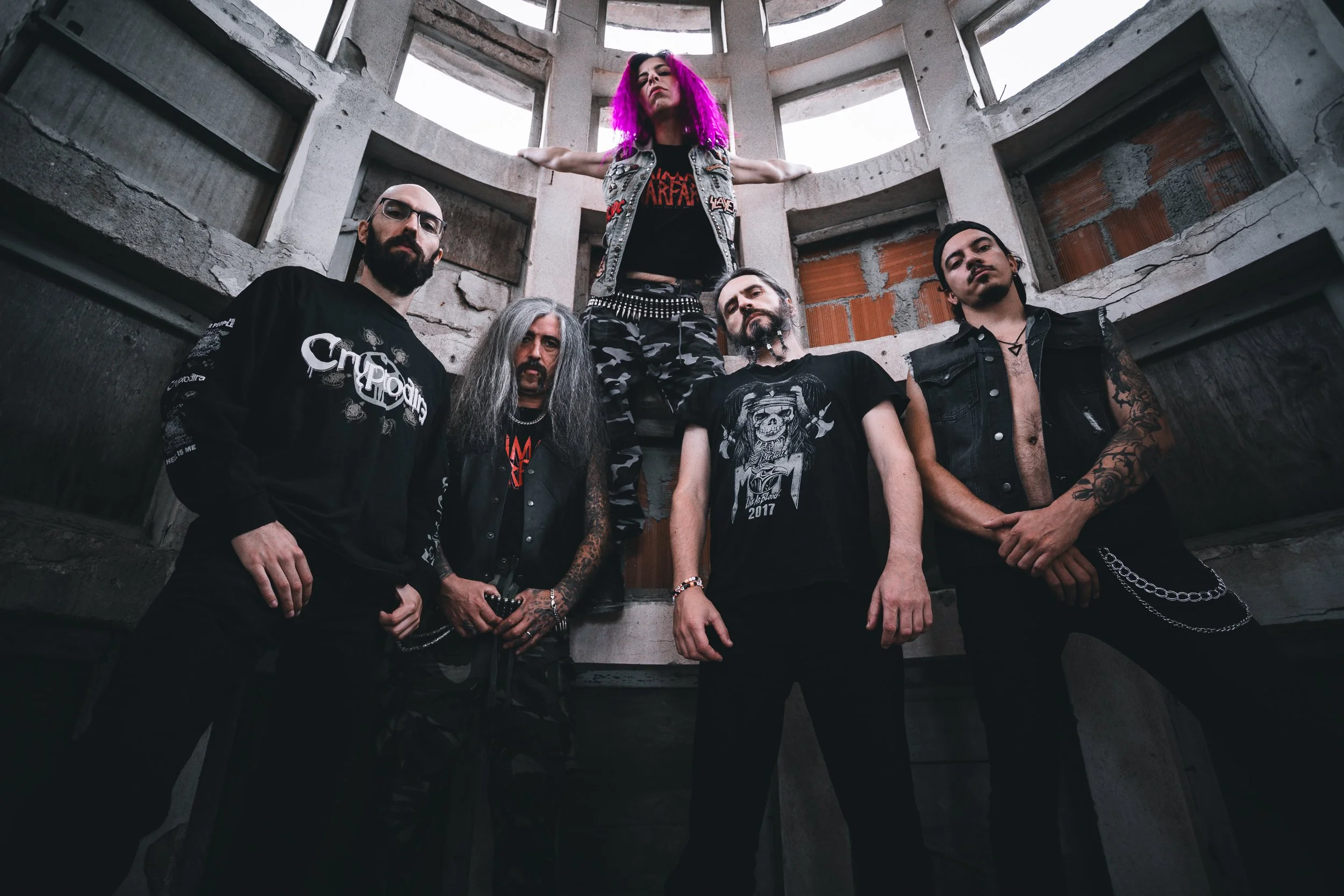 Grupo de cinco pessoas em ambiente industrial, com uma pessoa ao centro de vestimenta preta e cabelo cinza, duas ao lado com roupas pretas e tatuagens visíveis, uma com cabelo longo preto e outra com cabelo curto e evidente. Uma pessoa está numa estr