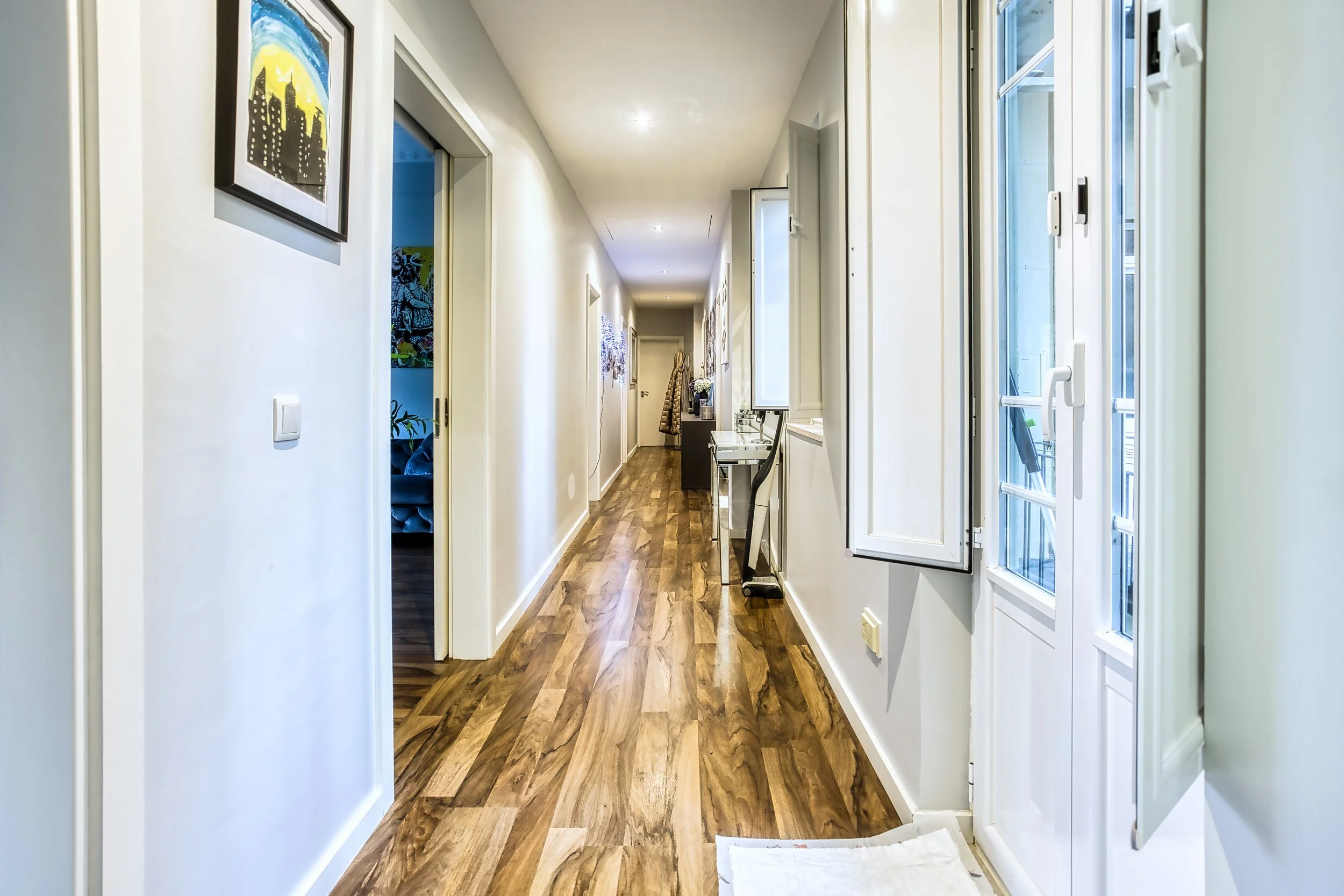 Corredor de casa com paredes brancas, piso de madeira e janelas abertas ao lado direito