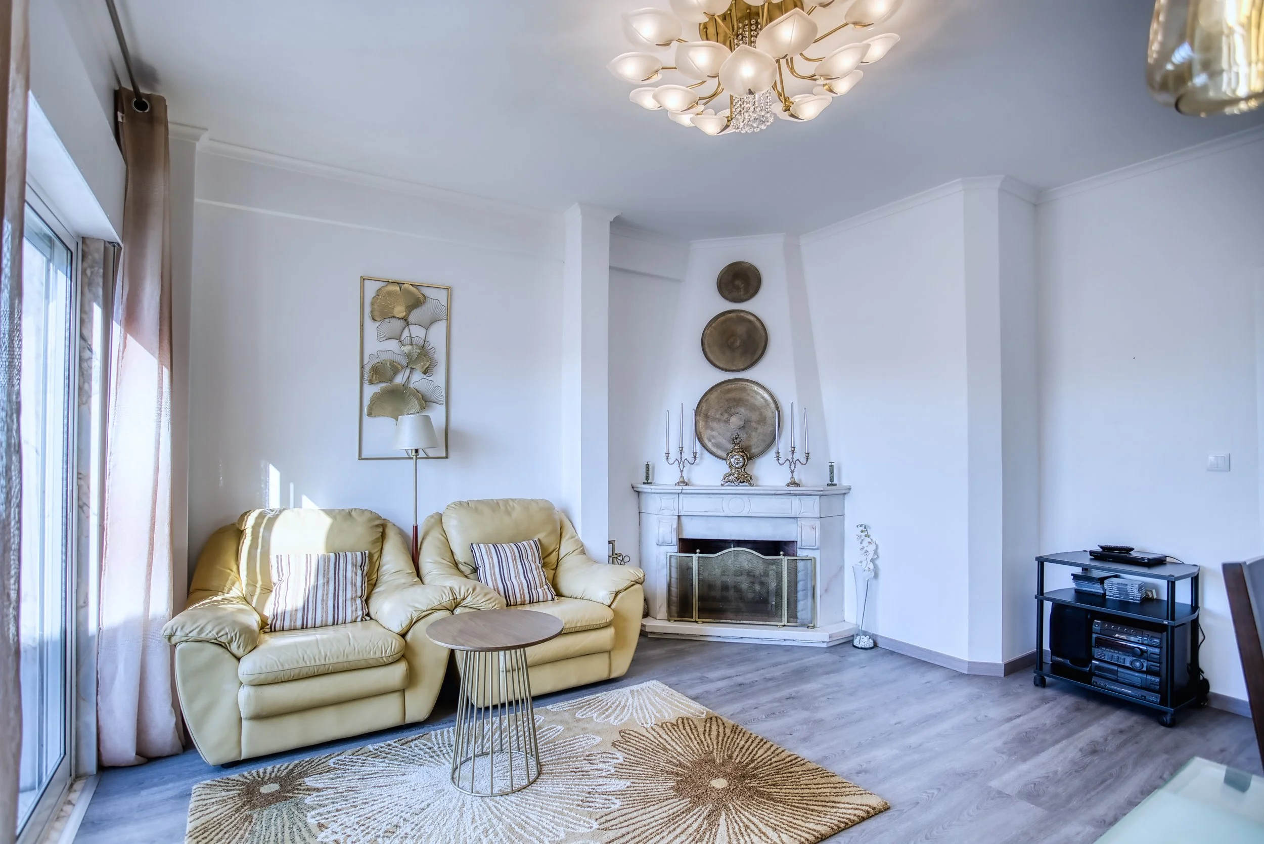 Sala de estar com sofá bege, mesa de centro redonda e tapete com padrão de sol. Parede branca com decoração de quadro de folhas douradas, lareira branca com três pratos decorativos acima, e canto com televisora preta sobre rack com aparelho de som. J