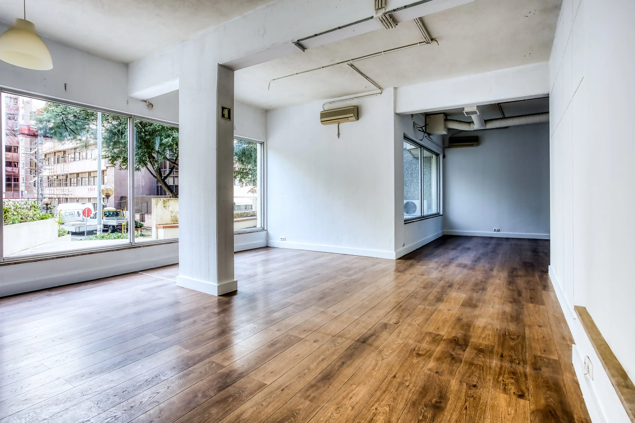 Sala vazia com janelas grandes, piso de madeira, paredes brancas, ar-condicionado e iluminação natural