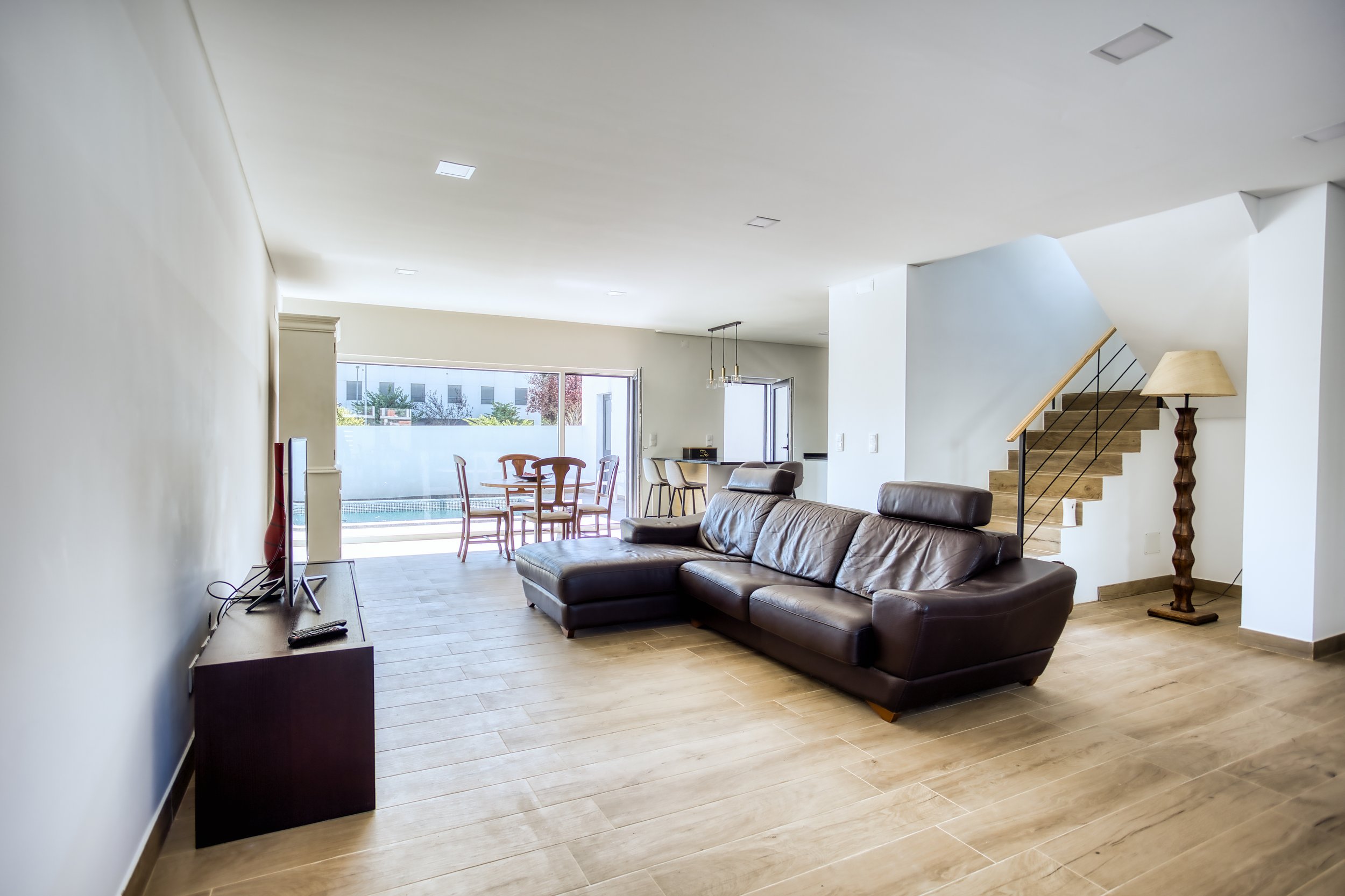 Sala de estar com sofá de couro marrom, televisão na parede, piso de madeira clara e parede branca. Ao fundo, uma mesa de jantar com cadeiras próximas a uma janela grande, com uma área de cozinha ao lado. Escada com degraus de madeira e corrimão de m
