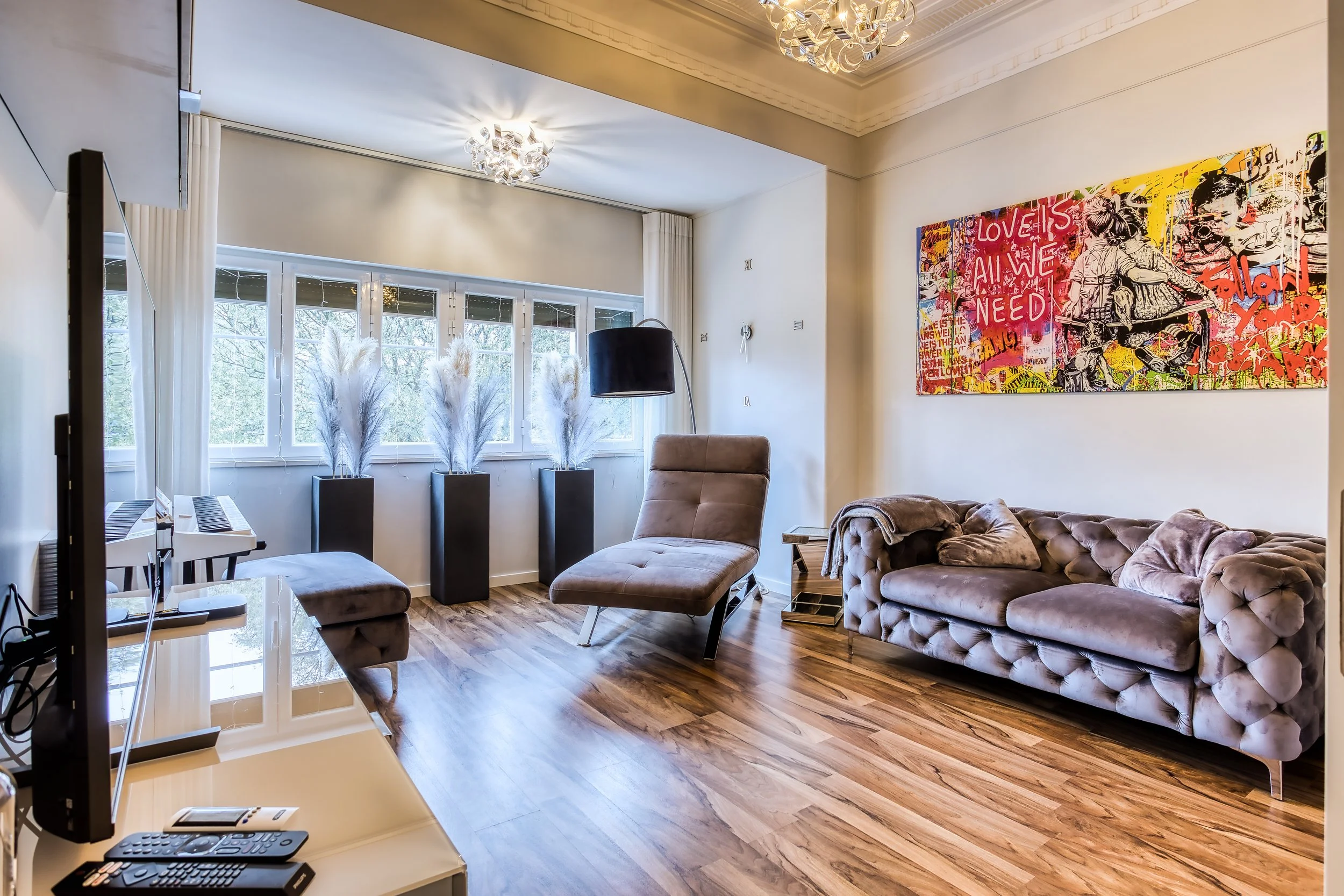 Sala de estar com sofá em veludo, poltrona reclinável, mesa lateral, quadro de arte colorida na parede, piso de madeira, janelas grandes com cortinas brancas, e decoração moderna.