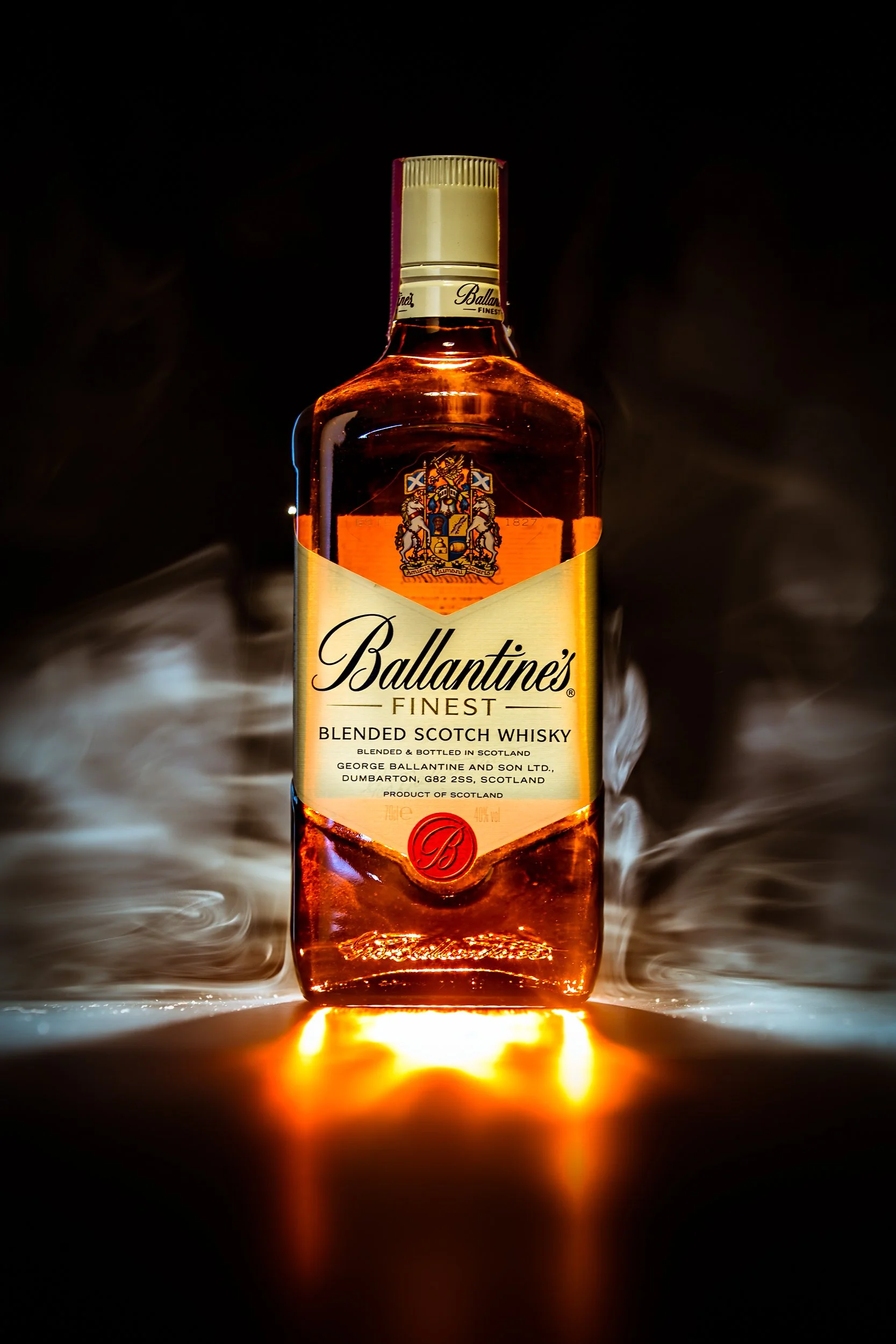 Garrafa de uísque escocês bolinha, marca Ballantine's, com fundo preto, iluminação destacando a garrafa e fumaça ao redor.