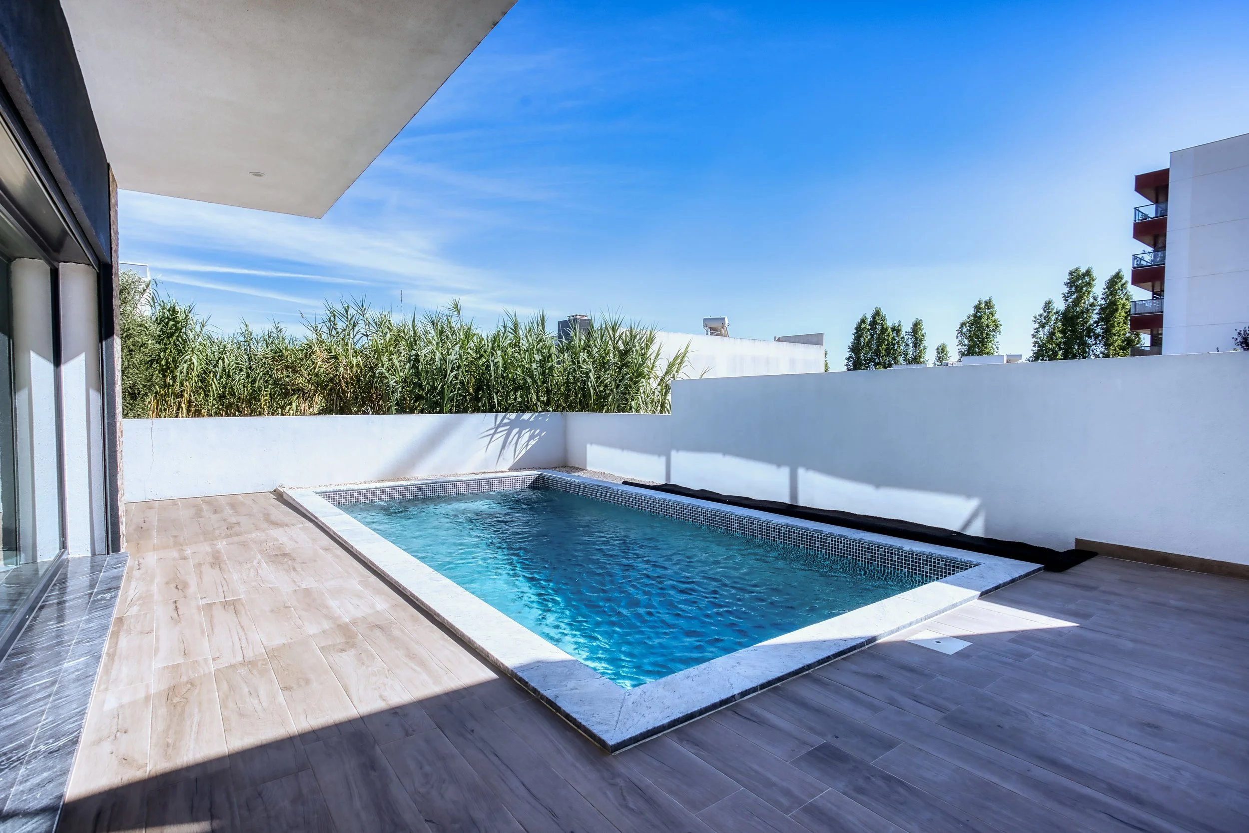 Varanda com piscina, pisos de madeira clara, muro branco, vegetação ao fundo e prédio alto à direita, sob céu azul com algumas nuvens.
