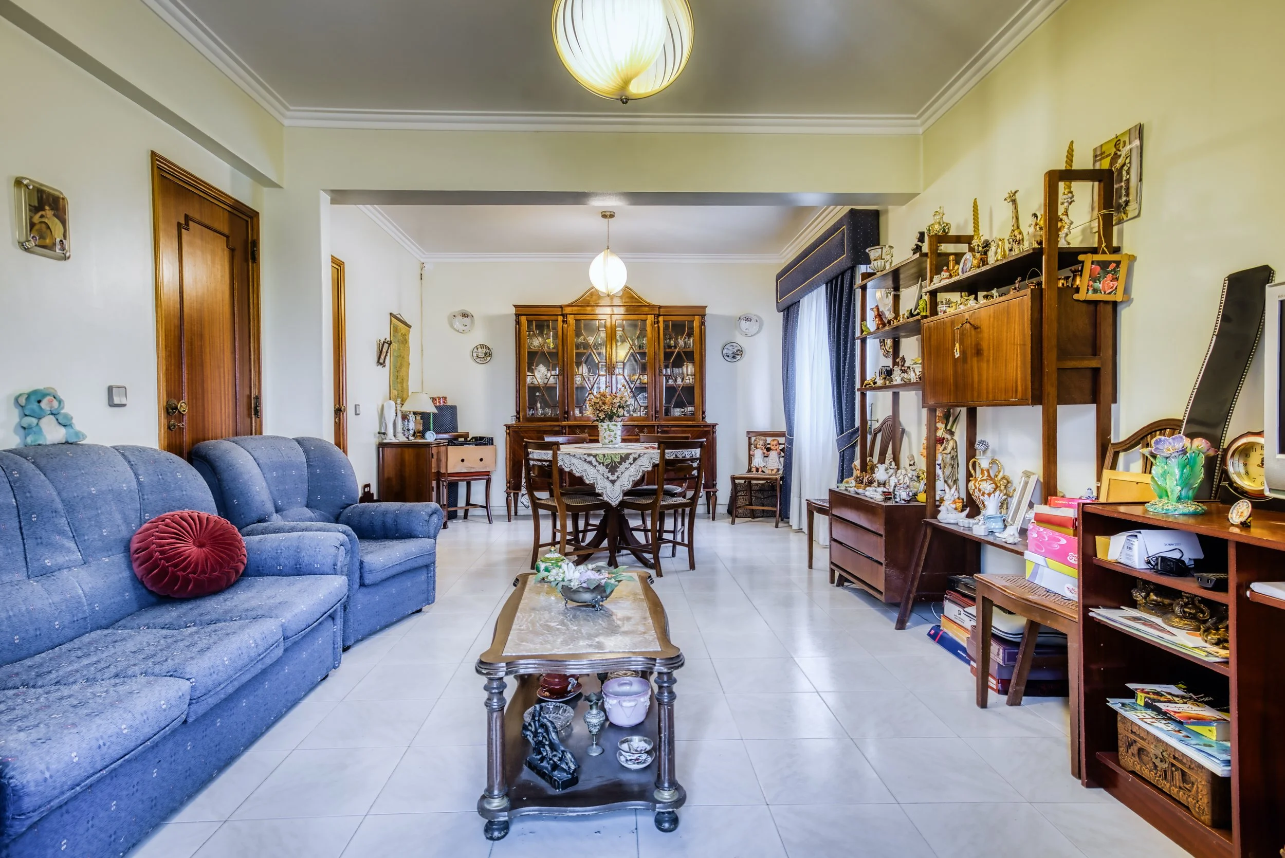 Sala de estar decorada com móveis de madeira e objetos de decoração, incluindo um sofá azul com almofada vermelha e uma mesa de centro, uma cristaleira de madeira, prateleiras com objetos e uma mesa de jantar com cadeiras. Há cortinas azuis na janela