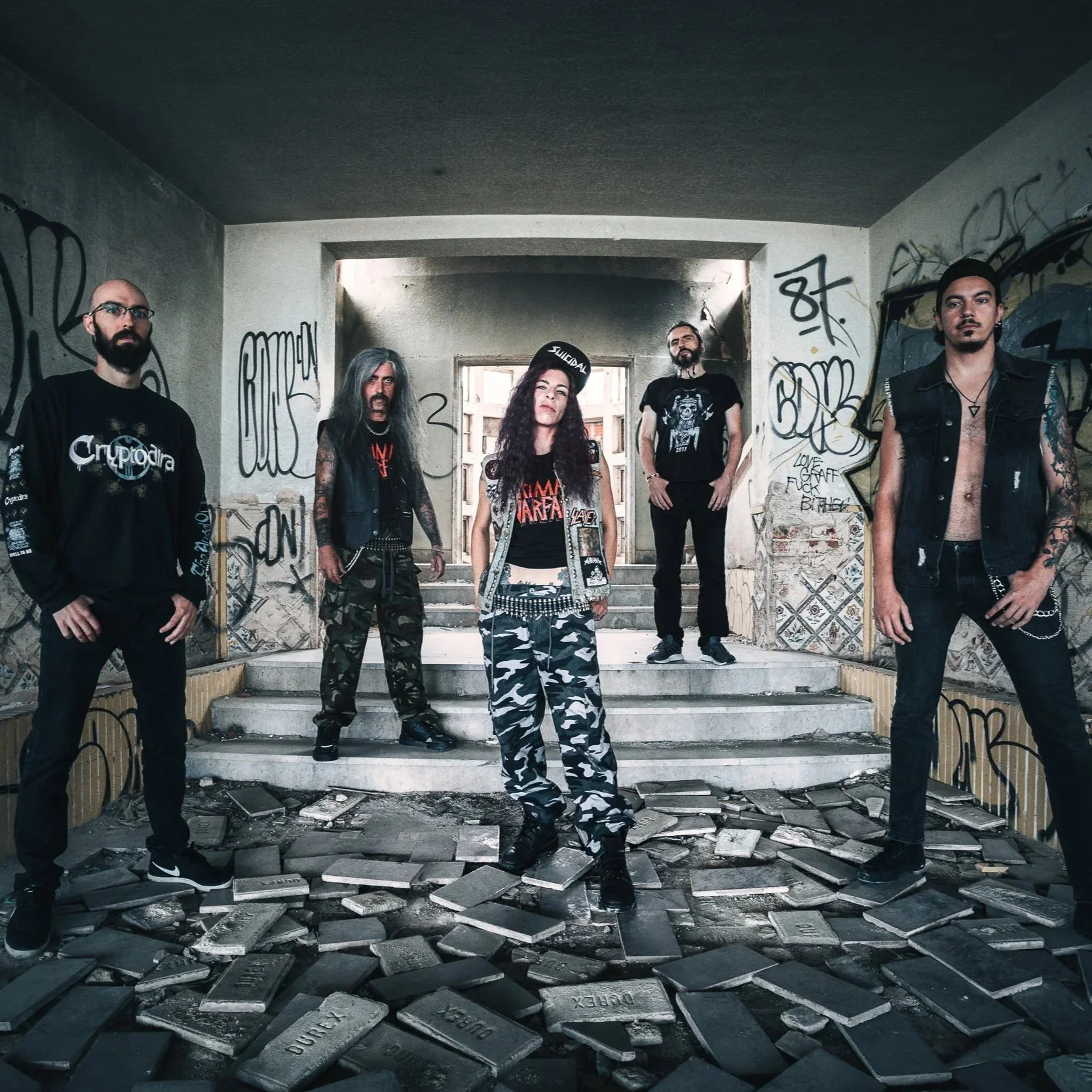 Cinco pessoas com roupas de estilo punk ou rock, em um ambiente com paredes grafitadas e chão com blocos de metal, posando para uma foto de grupo.