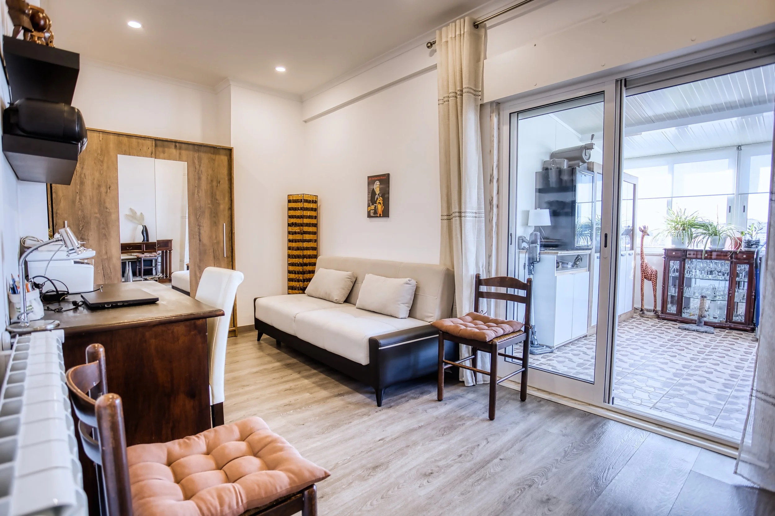 Sala de estar com sofá branco, poltrona, mesa de trabalho com laptop, e varanda com plantas e uma girafa de pelúcia ao fundo.