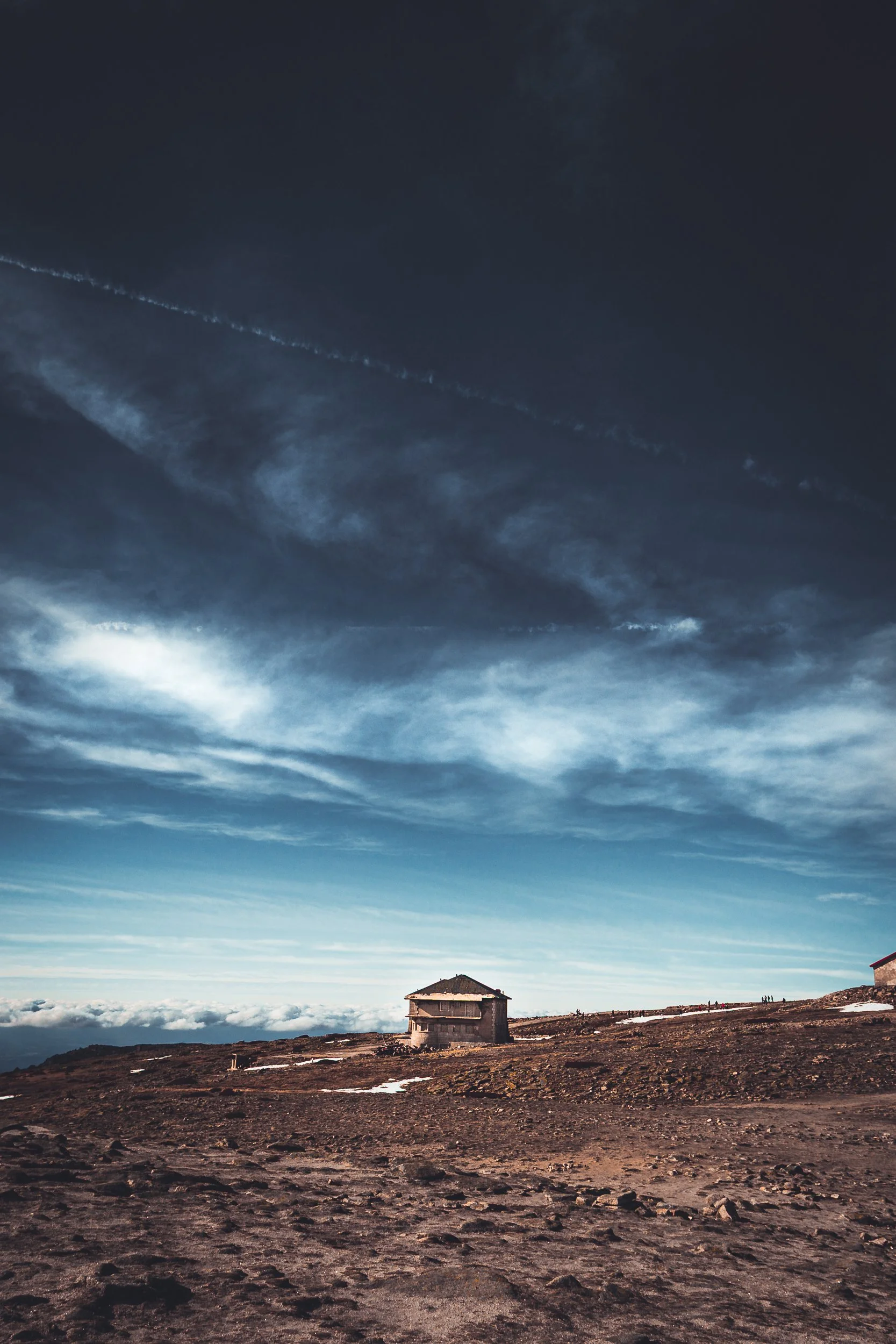 Cena de uma paisagem de montanha com um céu dramático e uma casa solitária na encosta, com nuvens ao fundo.