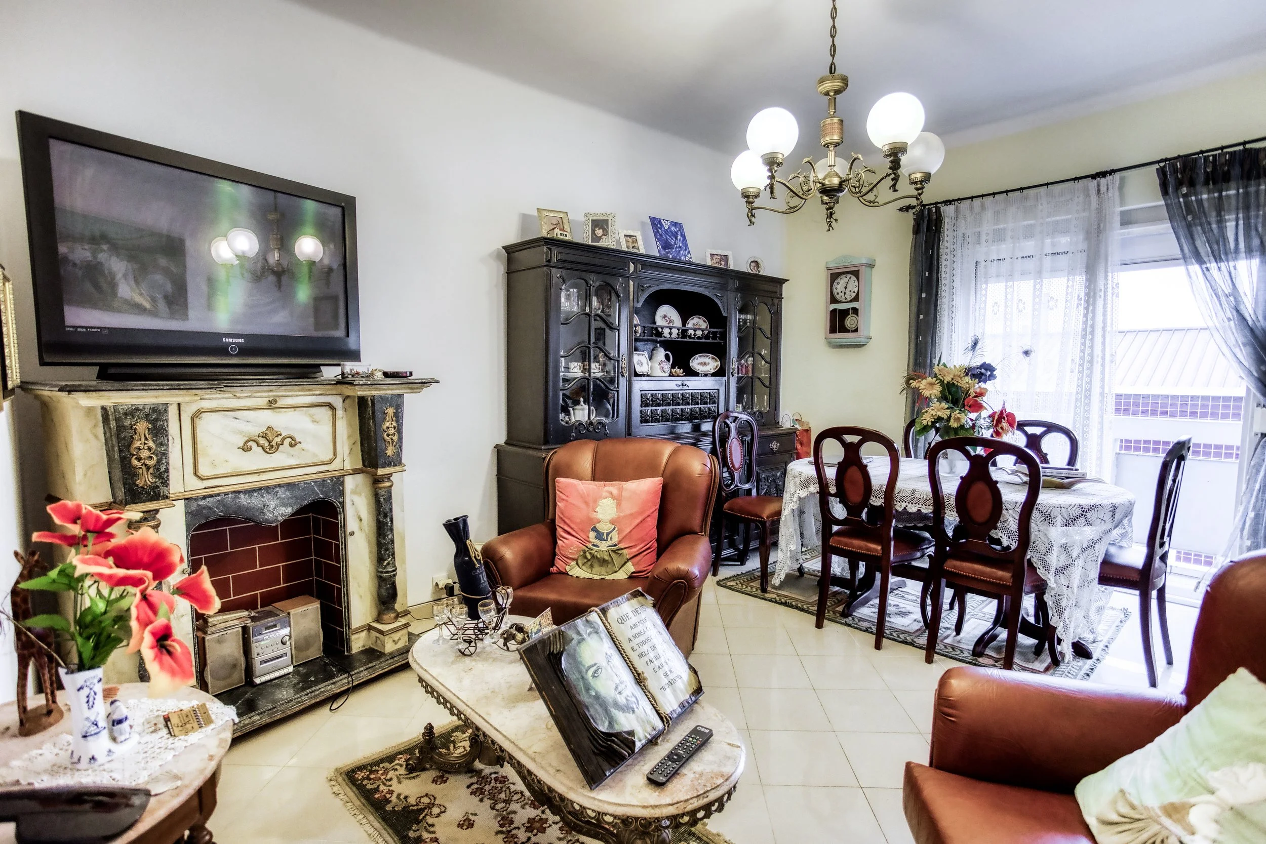 Sala de estar com sofá de couro, poltrona, mesa de centro, lareira decorativa, televisão, estante de madeira com louças, mesa de jantar com toalha de renda, cadeiras, janela com cortinas de tecido, luminária de teto com luzes brancas.