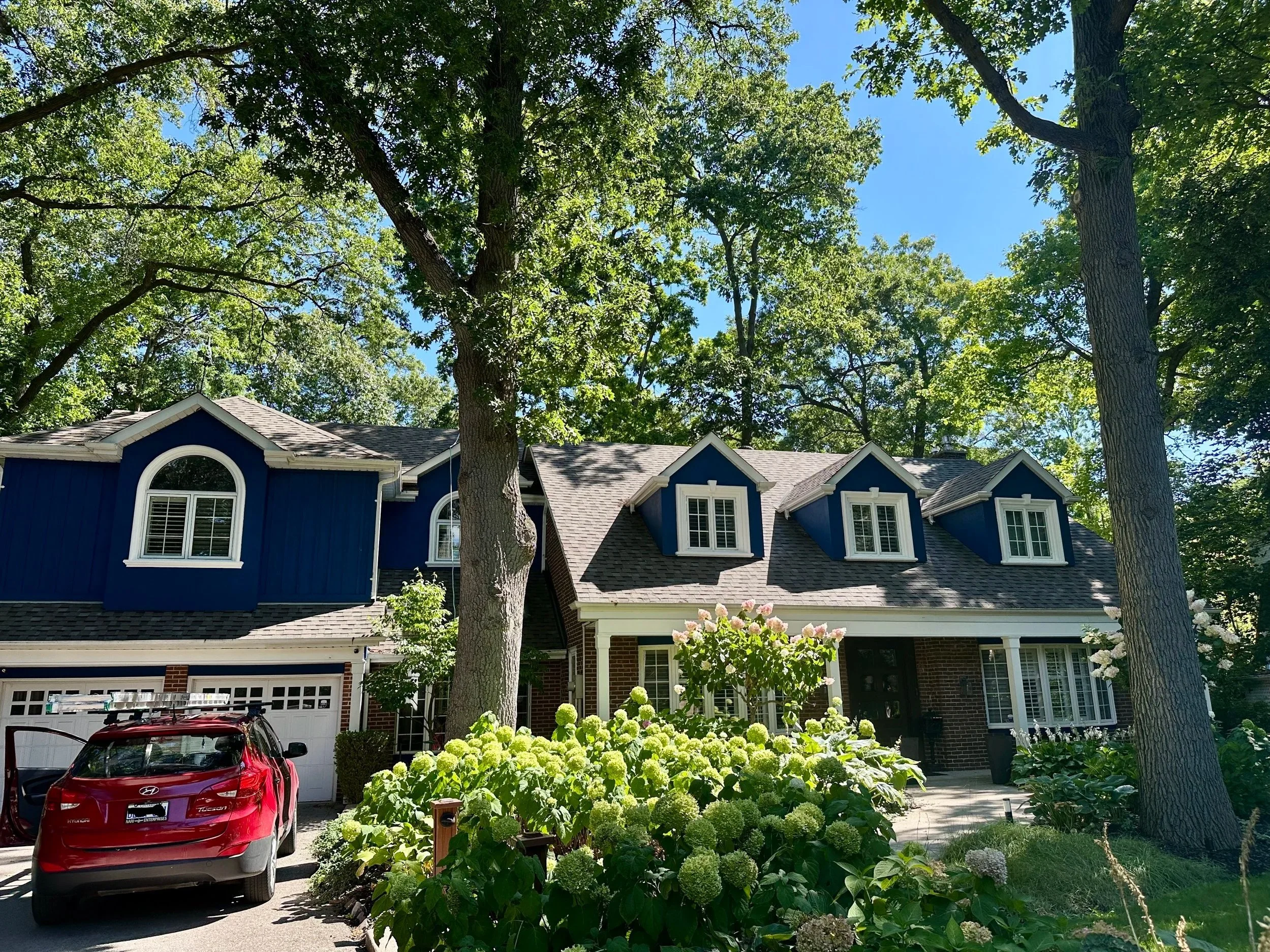 Front view of a house with blue siding and dormer windows, surrounded by trees and blooming bushes, with a red car parked in driveway.