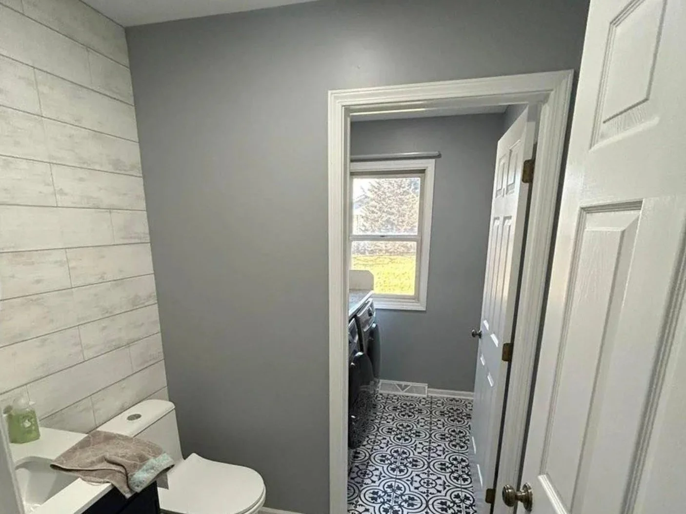 Bathroom with a toilet and a small laundry area visible through an open door, with a window showing outside greenery.