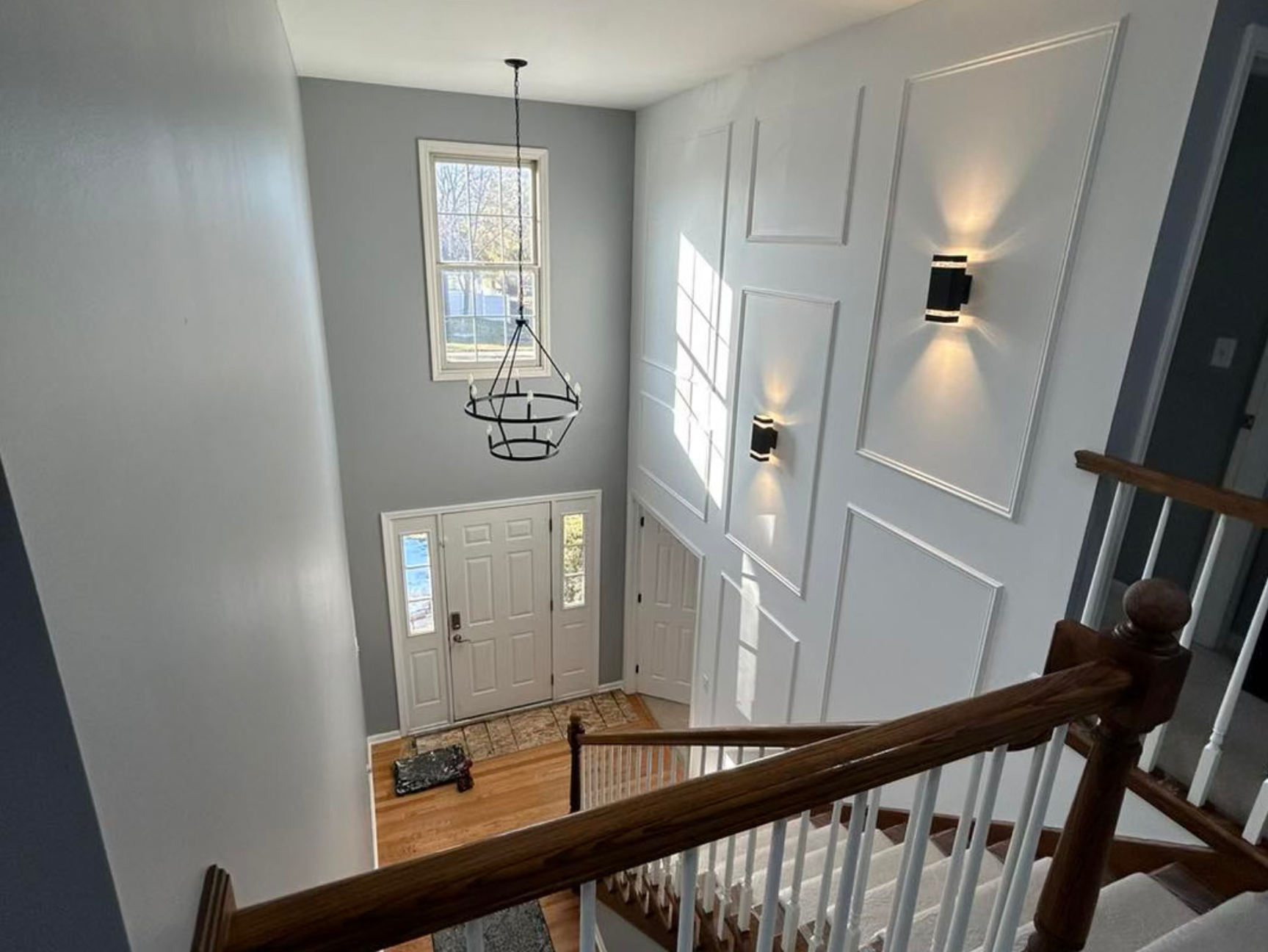 Indoor view of a modern front entryway with double doors, a staircase with wooden handrails, wall-mounted lights, and a large window allowing natural light.