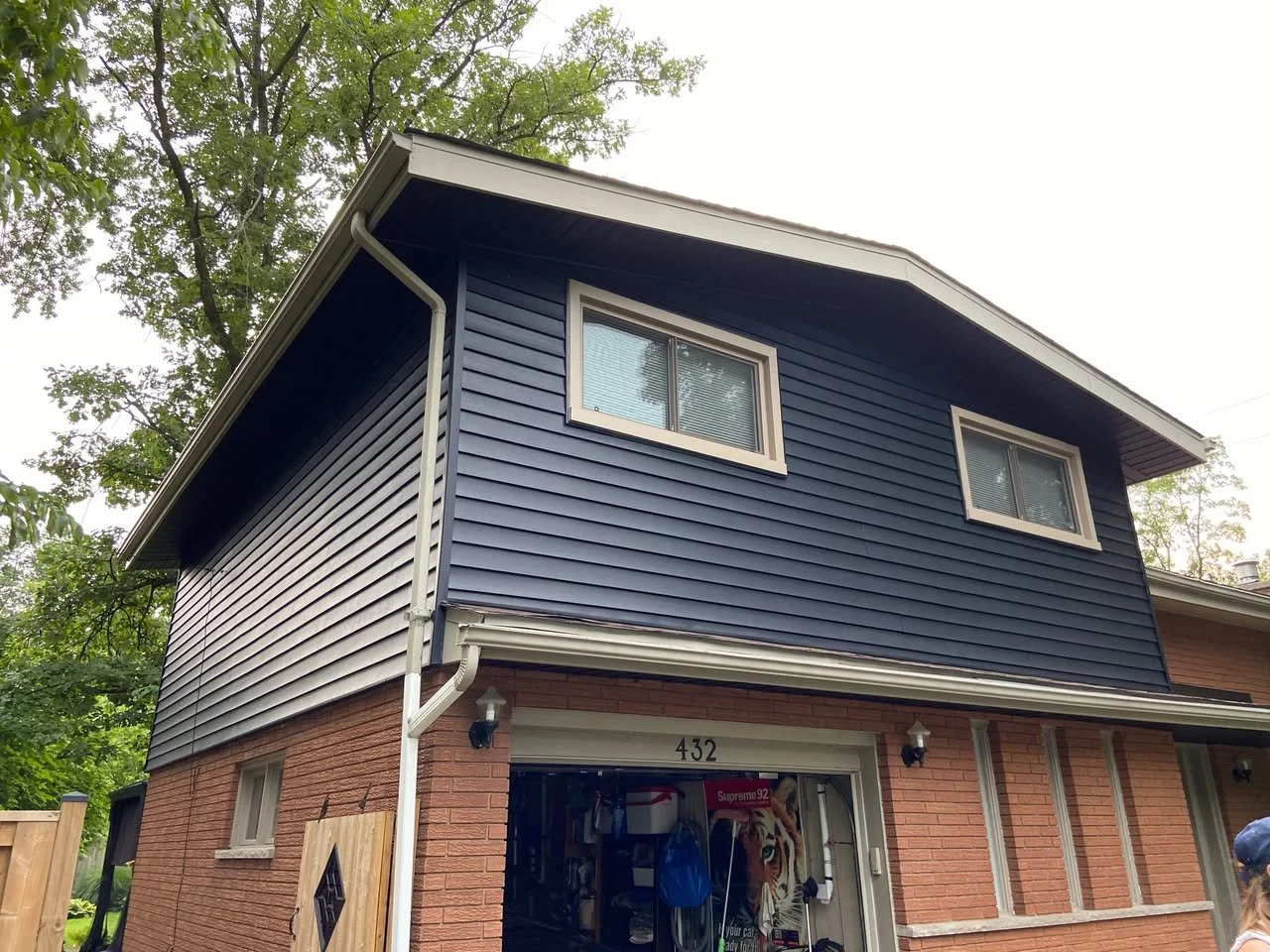 Two-story house with brick lower level and blue siding upper level, with two windows on the upper floor, garage door open showing a dog poster and various items inside, house number 432, surrounded by trees.