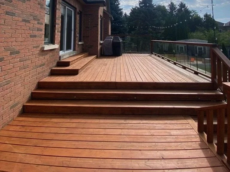 Newly built wooden deck with steps leading up to it, attached to a brick house, with a glass railing on one side and outdoor furniture and trees in the background.
