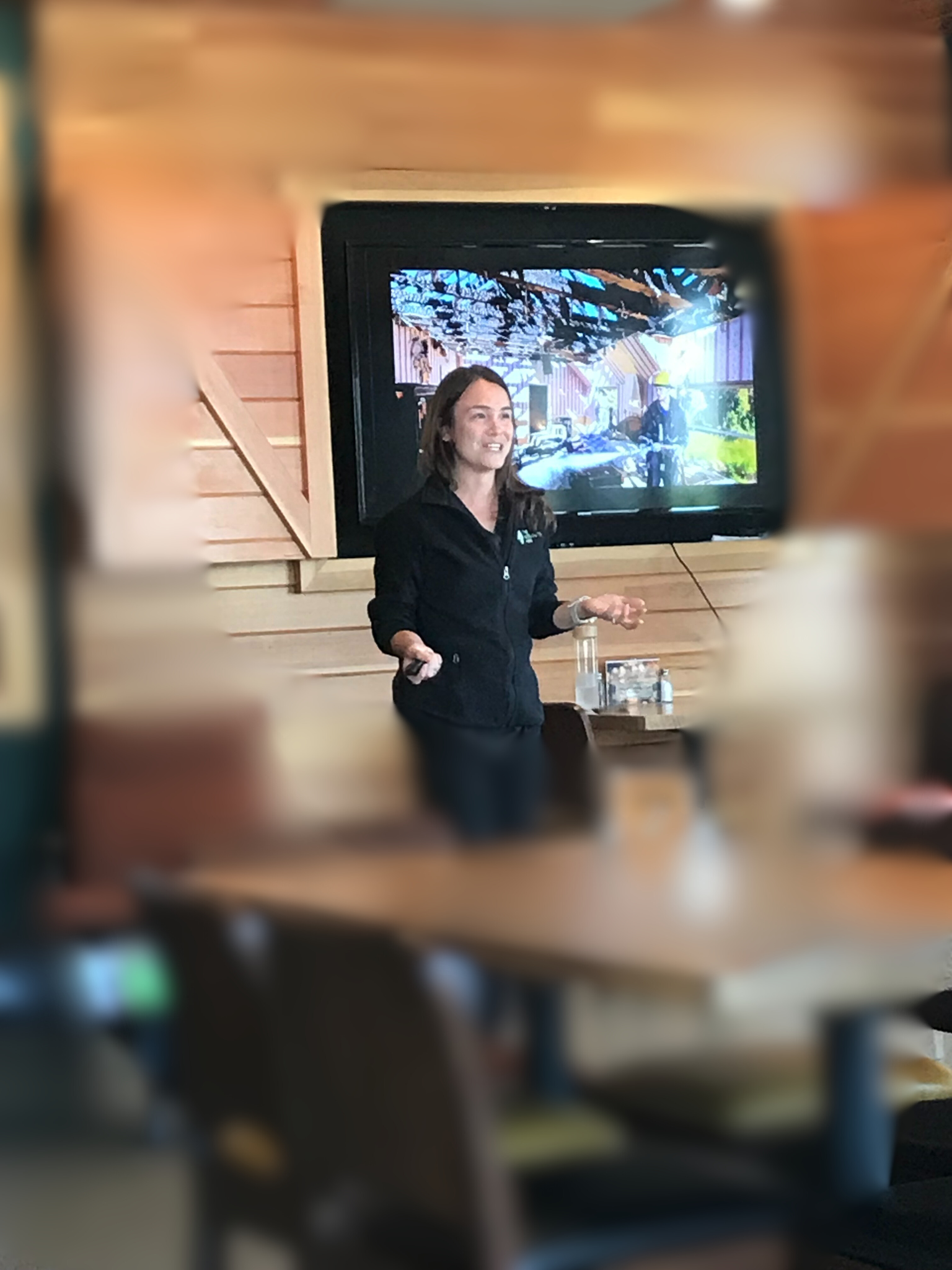 A woman giving a presentation in front of a large screen in a wooden-paneled room.