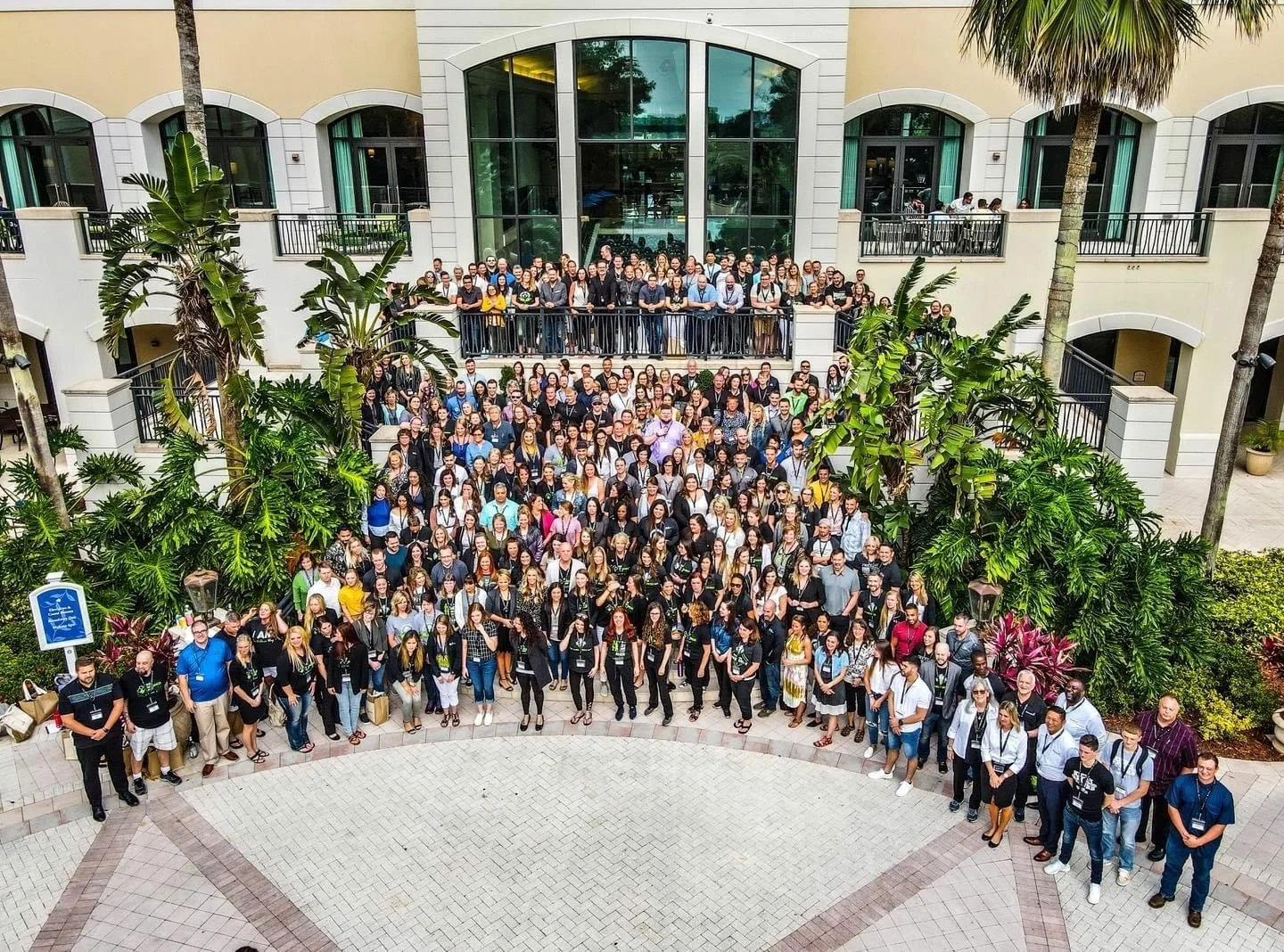 A large group of people gathered together outdoors for a group photo, standing on a paved area with tropical plants and trees in a courtyard of a multi-story building with balconies.