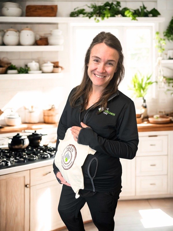 A woman with long brown hair, smiling and dressed in black, holding a cloth bag with a 100% biodegradable eco-friendly logo in a bright kitchen with white cabinets and shelves, and green plants.