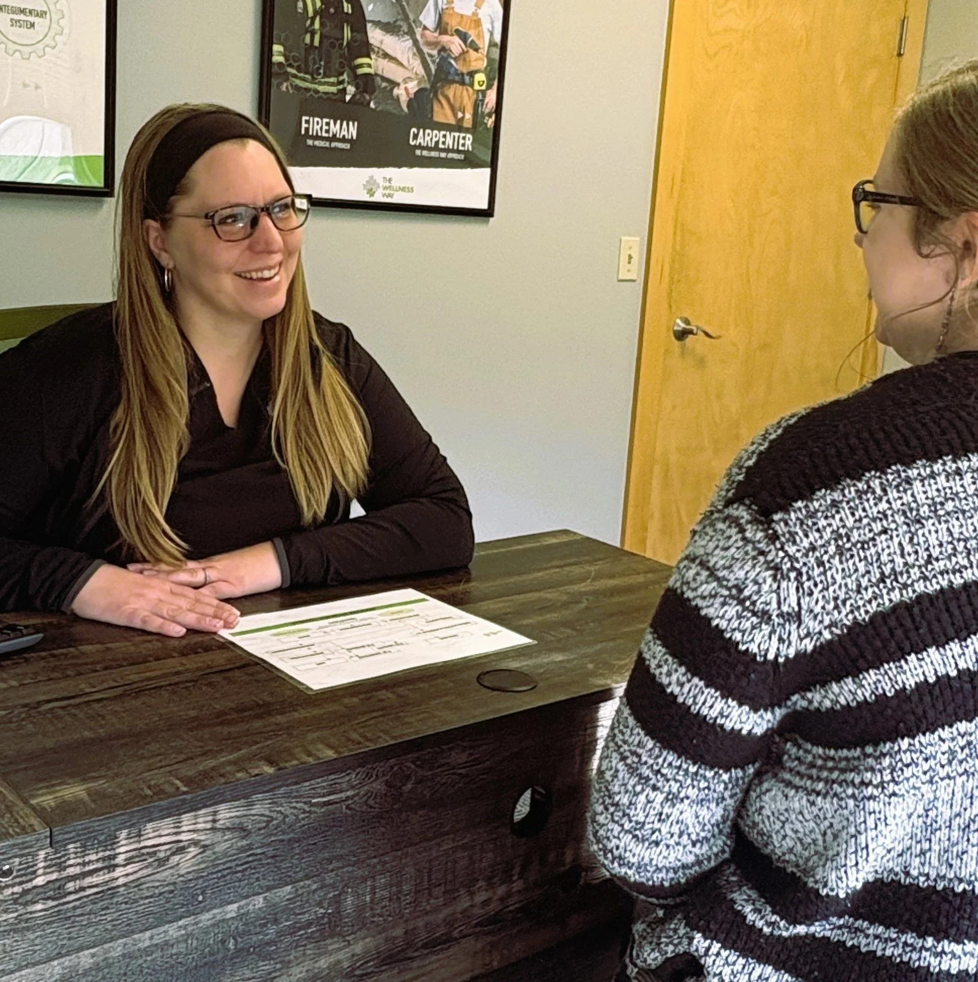 Two women engaged in conversation at a wooden desk in an office, with framed posters on the wall behind them.