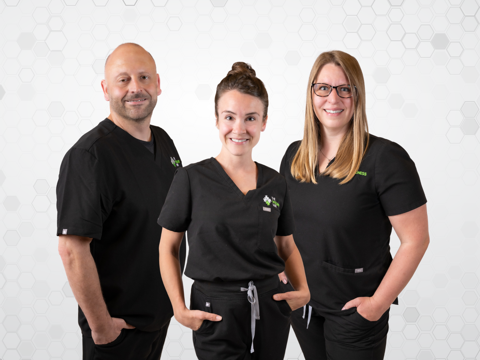 Two health coaches and a chiropractor standing together, smiling, wearing black scrubs against a white background with light gray hexagon pattern.