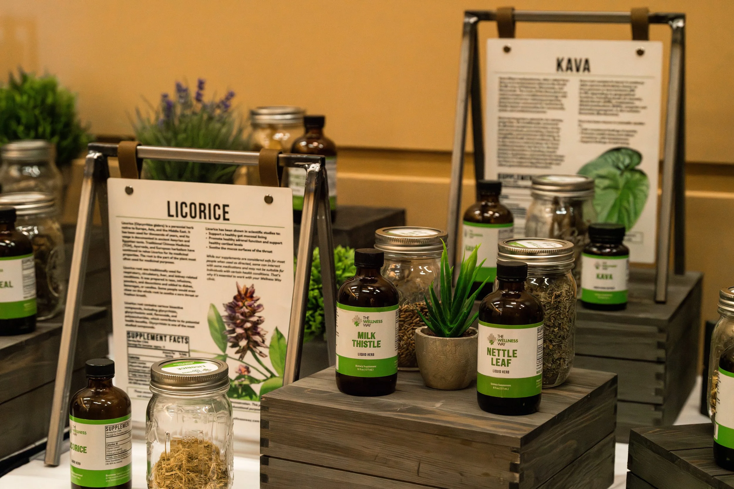 Display of herbal products, including bottles labeled "Milk Thistle" and "Nettle Leaf," jars of dried herbs, and informational signs about licorice and kava on wooden tables at an herbal shop or market.