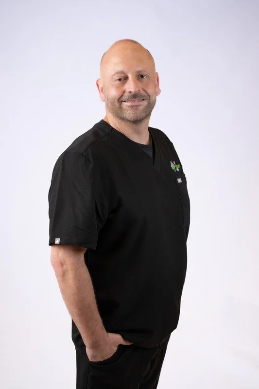A smiling man wearing a black scrub top, standing against a plain white background.
