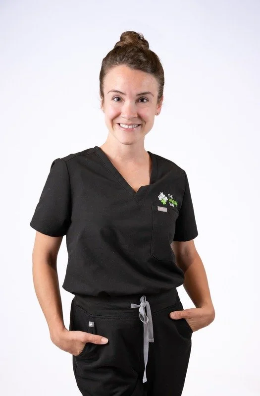 A woman with brown hair tied in a bun, wearing black scrubs with a logo, smiling at the camera against a plain white background.