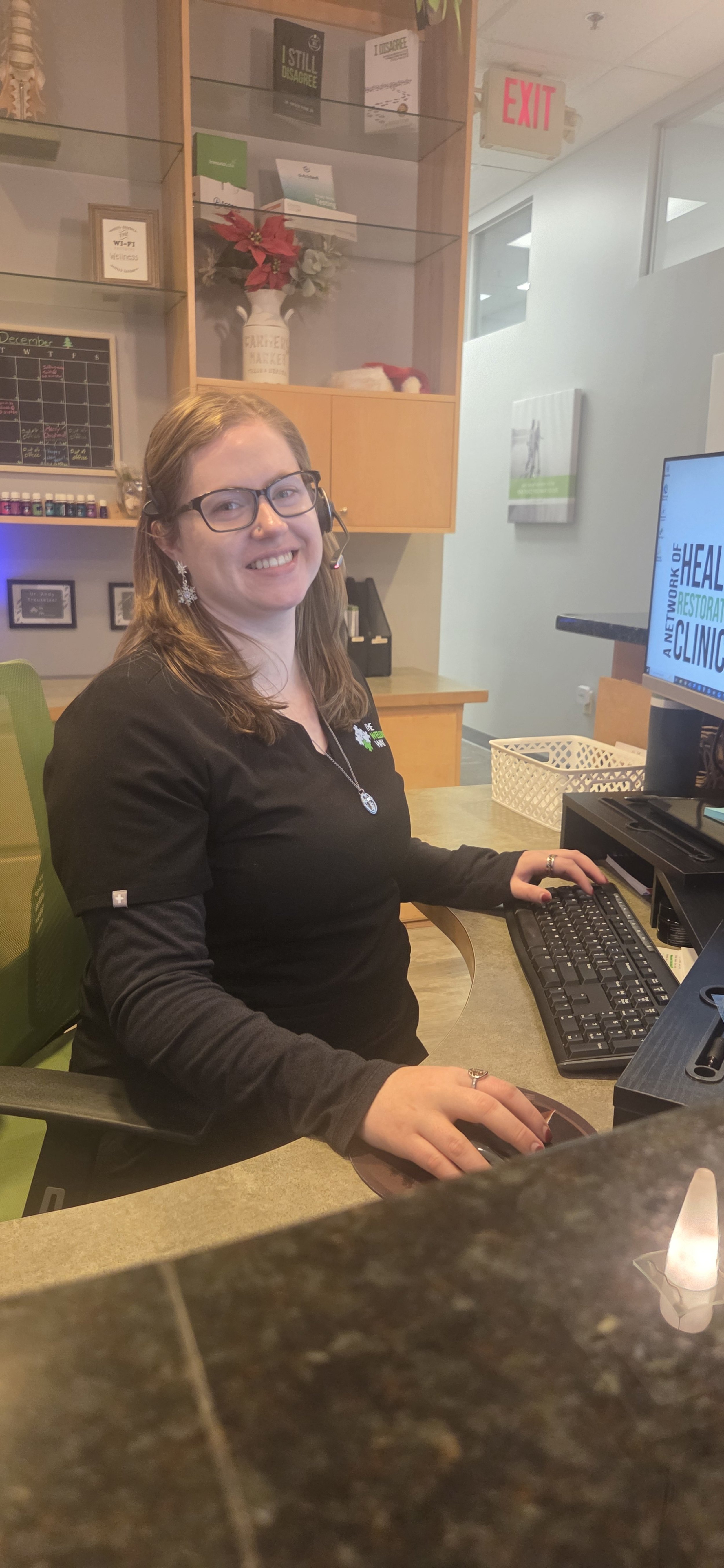 A woman with glasses and brown hair sitting at a desk, smiling at the camera, using a computer with a headset on, in an office or reception area.