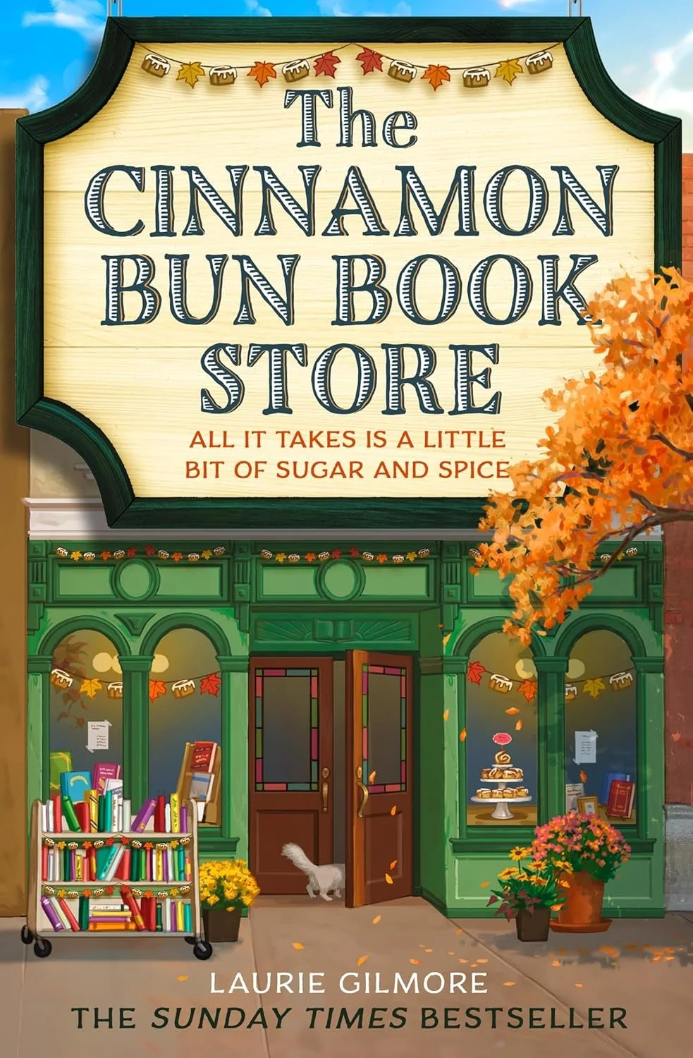Sign for The Cinnamon Bun Book Store with autumn decorations, bookstore window display, some books, a small table with a cake, and a white cat near the door.