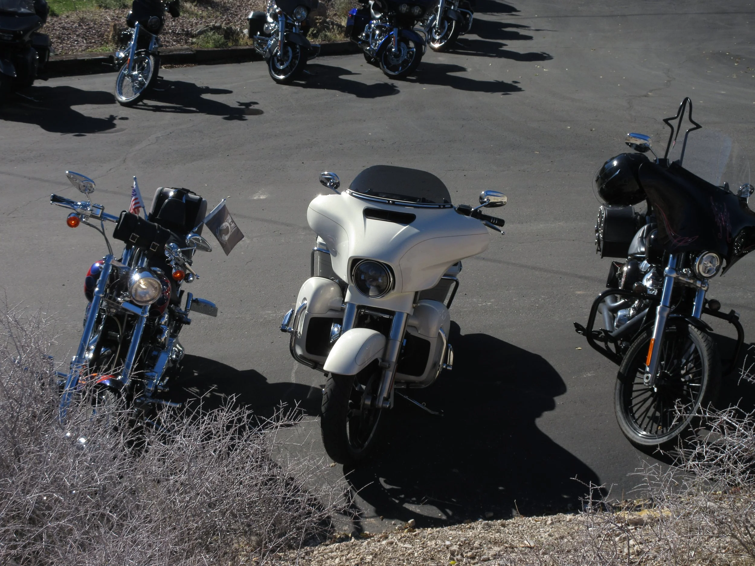 Three motorcycles parked on a paved area with a row of motorcycles in the background. The middle motorcycle is a white touring bike with a large front fairing, and the others are black cruisers with windshields and chrome details.