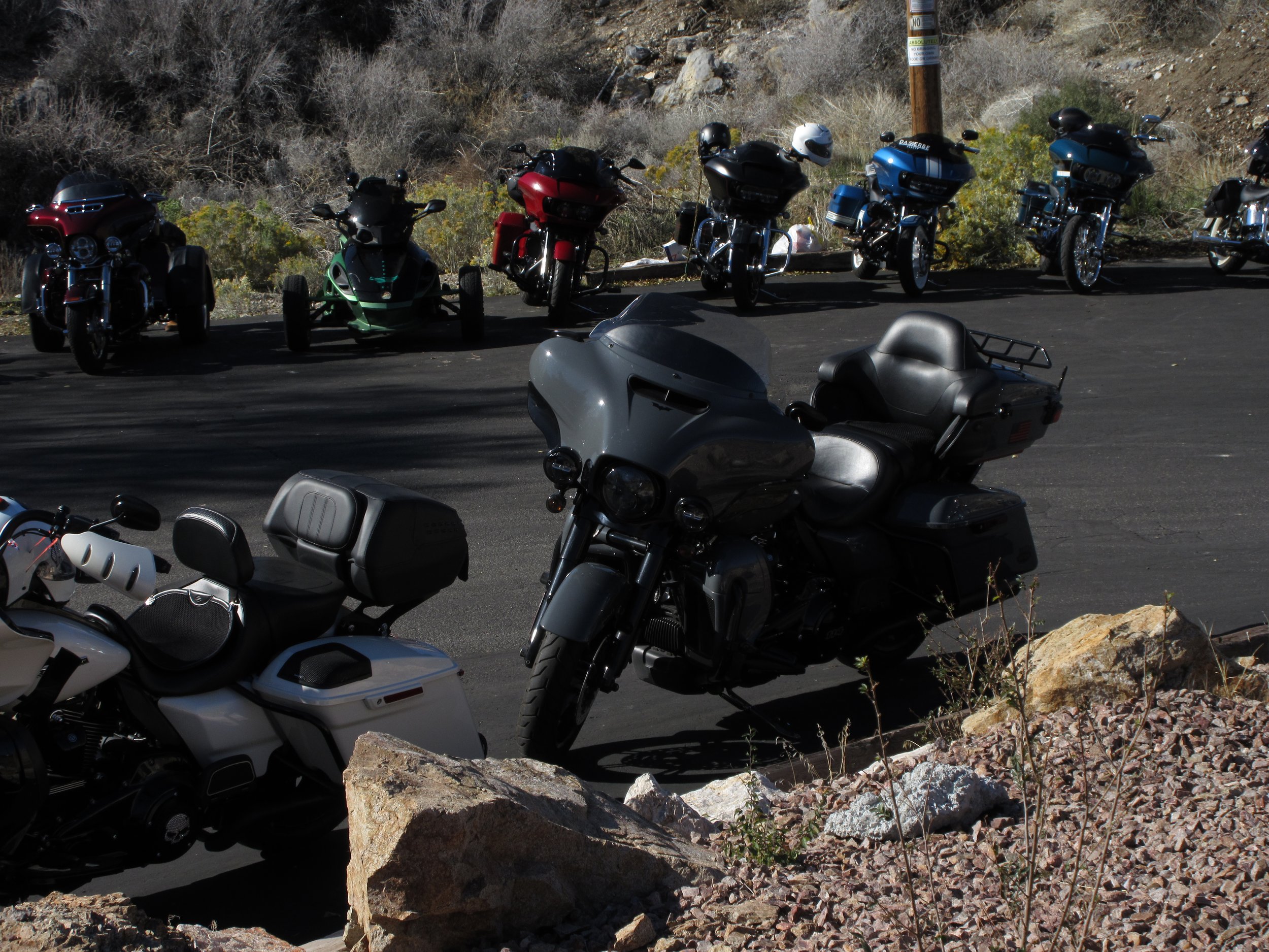 Group of parked motorcycles and three can-am three-wheeled motorcycles in a sunny outdoor area with rocky and shrub-covered terrain.