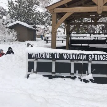 Snow-covered Welcome to Mountain Springs sign with trees and a wooden shelter in background.