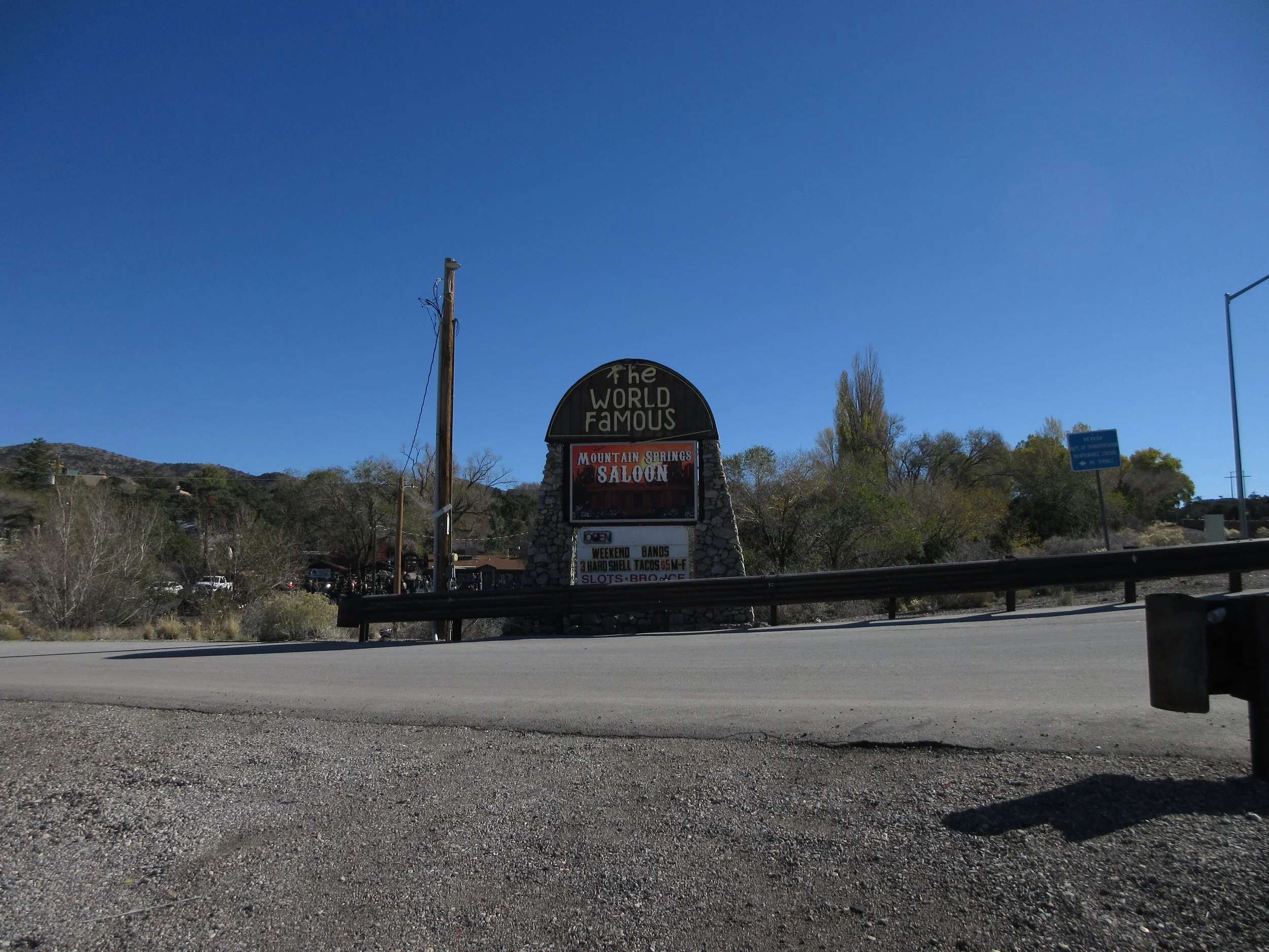 Signboard reading 'The World Famous Mountain Springs Saloon,' with additional details about weekend bands and food, situated by a highway with trees and mountains in the background under a clear blue sky.