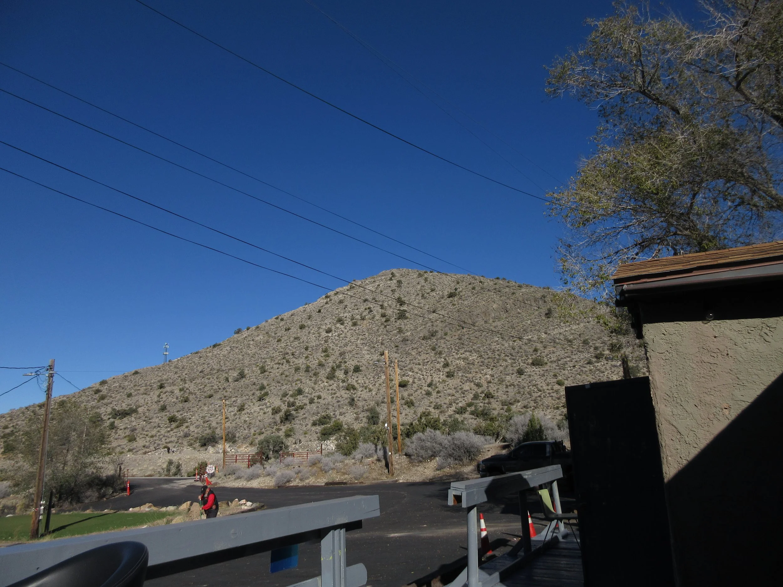 View of a hillside or mountain with sparse vegetation under a clear blue sky, with some power lines, trees, a building, and a person in the foreground.
