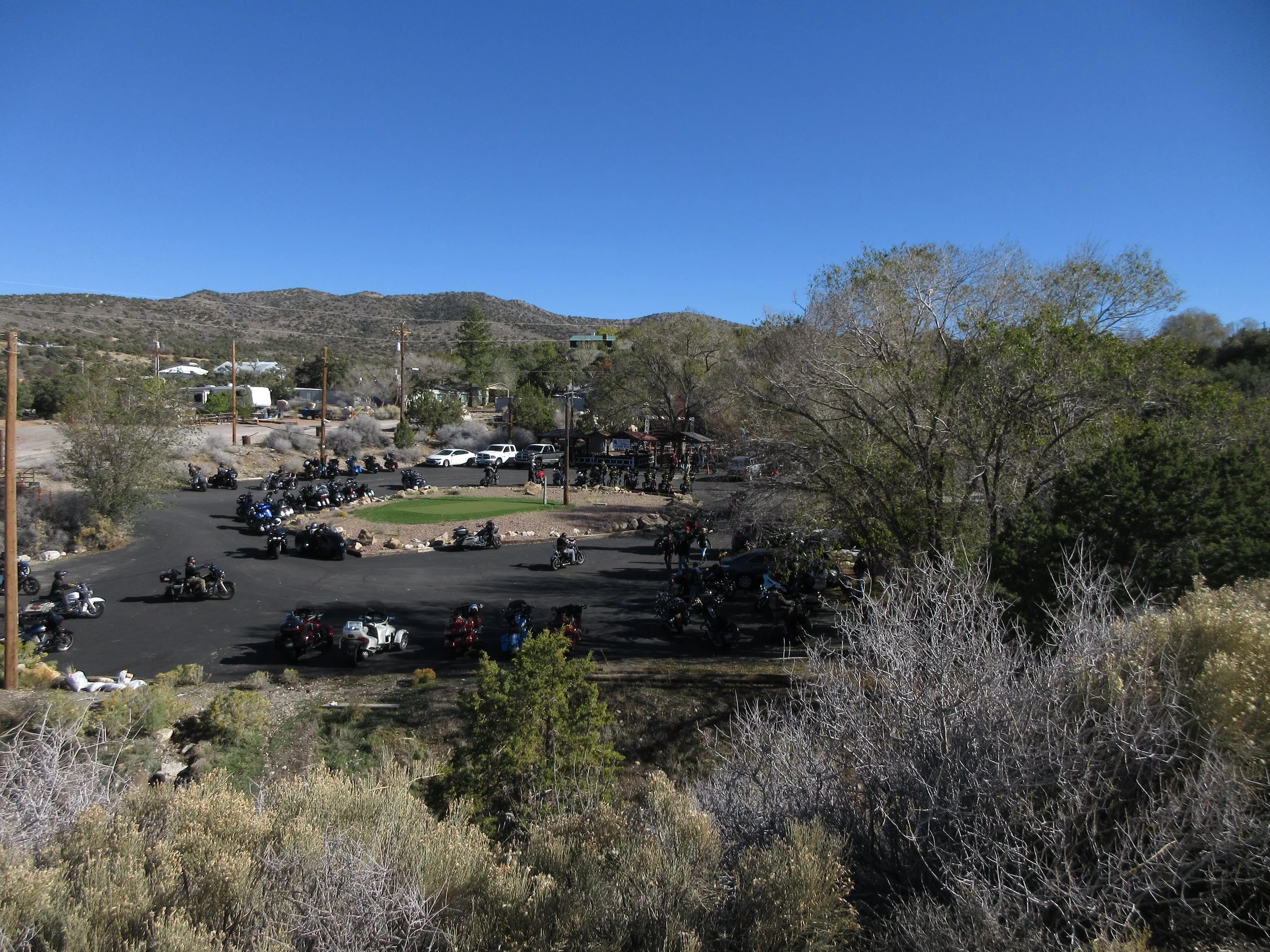 A parking lot filled with motorcycles and a few cars, surrounded by trees and shrubs, with hills and a clear blue sky in the background.