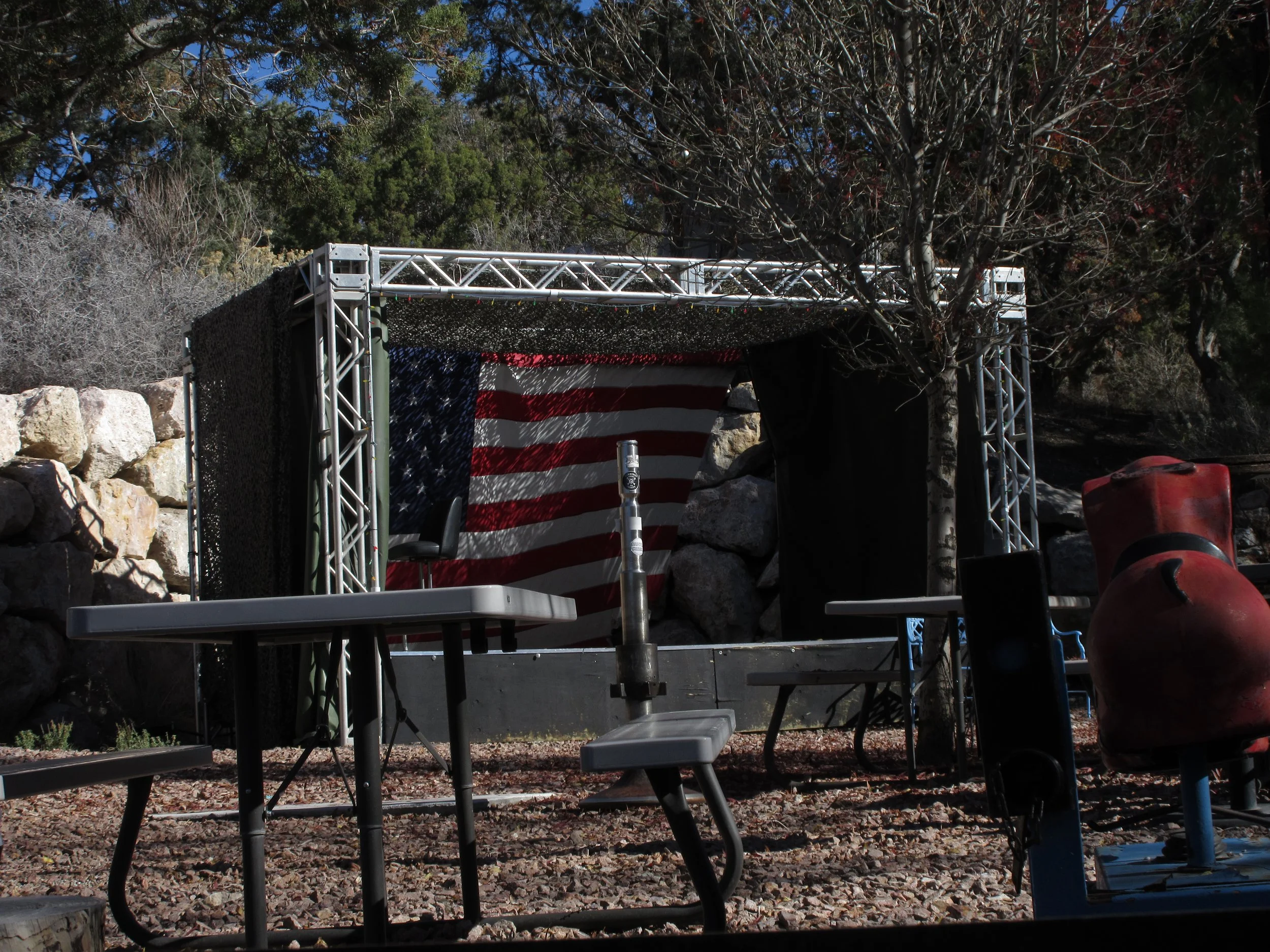 An outdoor stage with an American flag backdrop, surrounded by tables and chairs, trees, rocks, and a red inflatable figure.