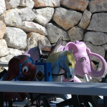 Collection of children's playground toys, including a pink elephant, a yellow horse, and a red figure, set on a metal surface against a stone wall background.