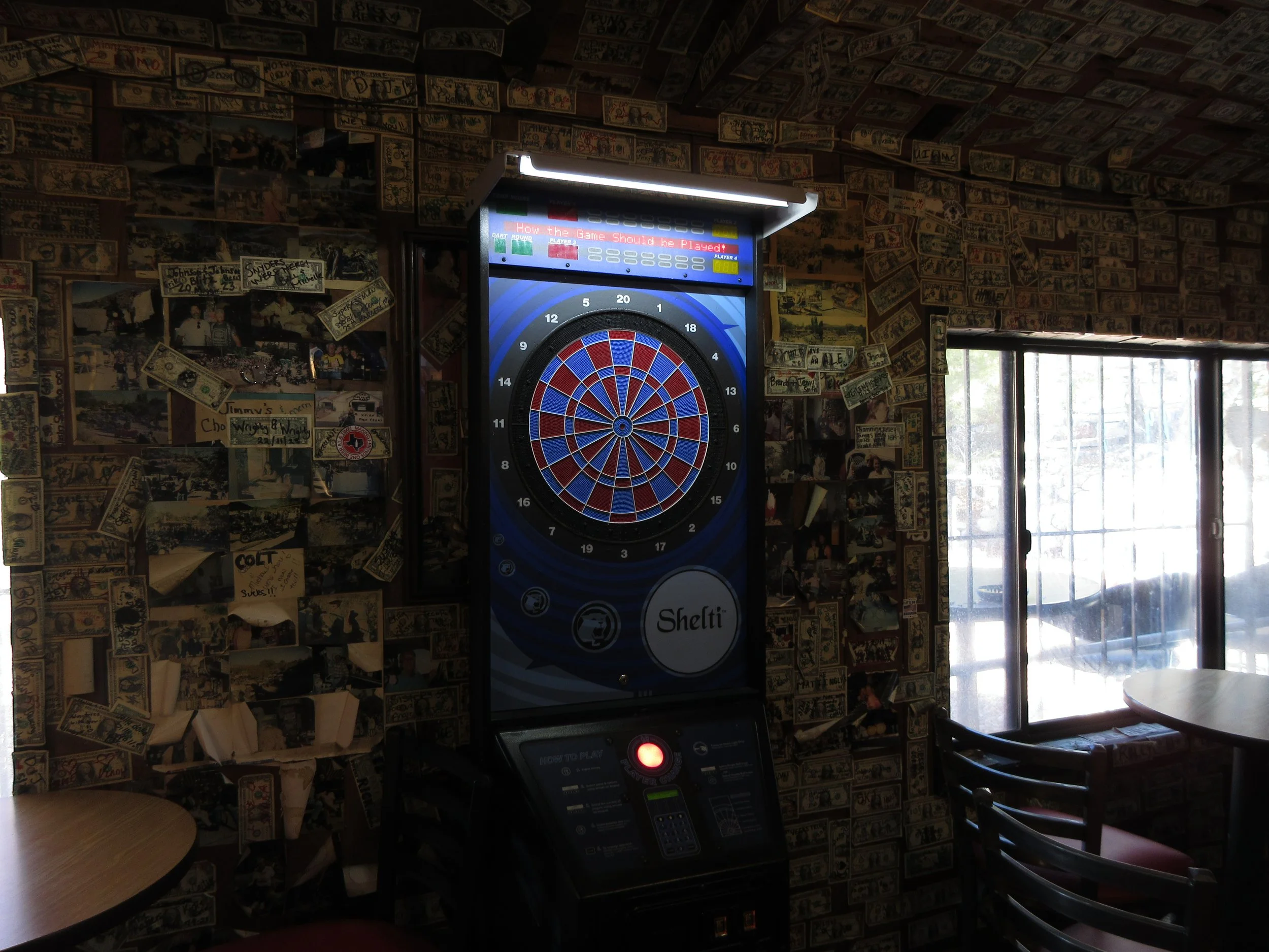 An electronic dartboard machine in a dimly lit room with walls decorated with dollar bills and photographs, near a window with natural light.